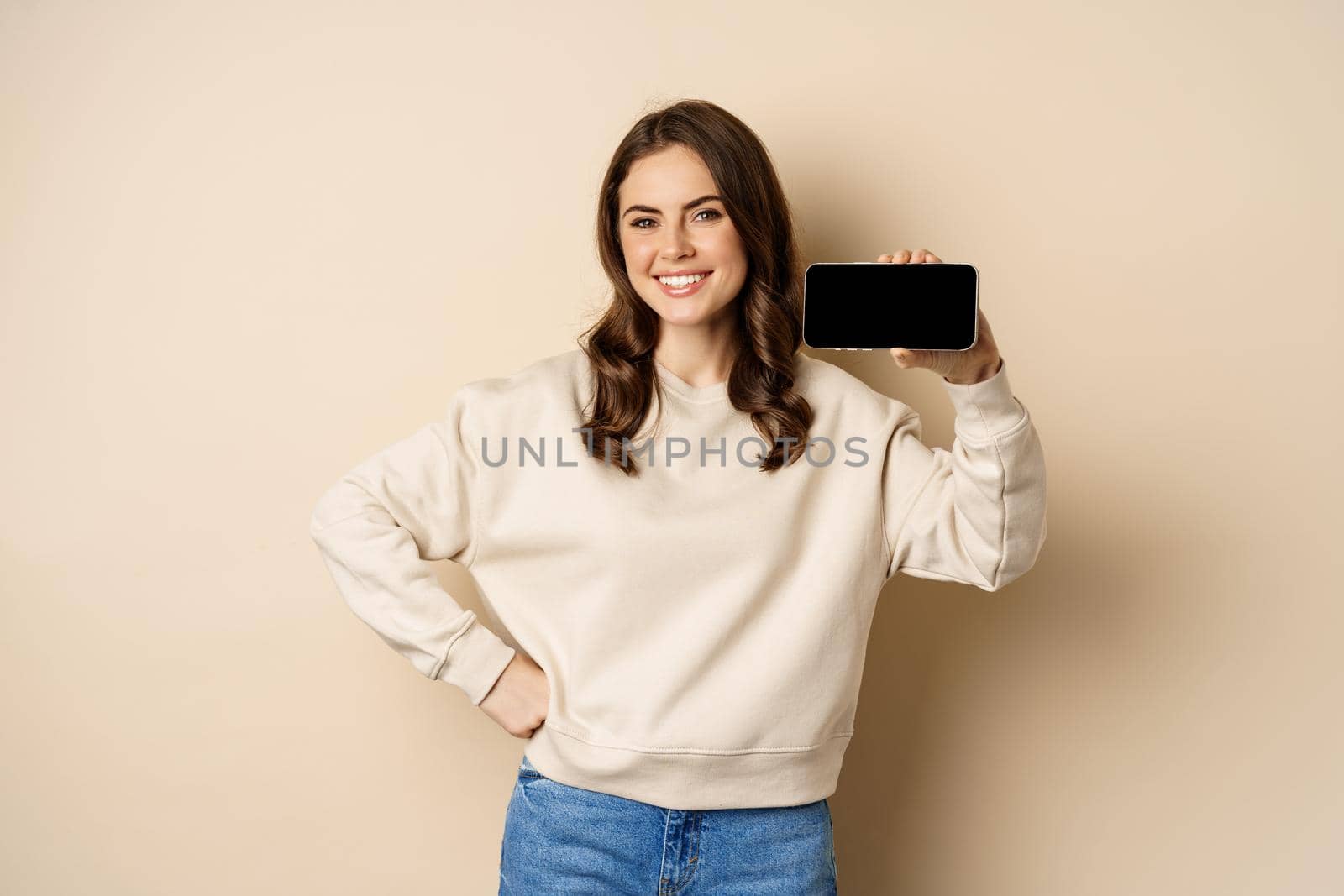 Portrait of smiling cute woman showing horizontal smartphone screen, website or application, standing over beige background by Benzoix