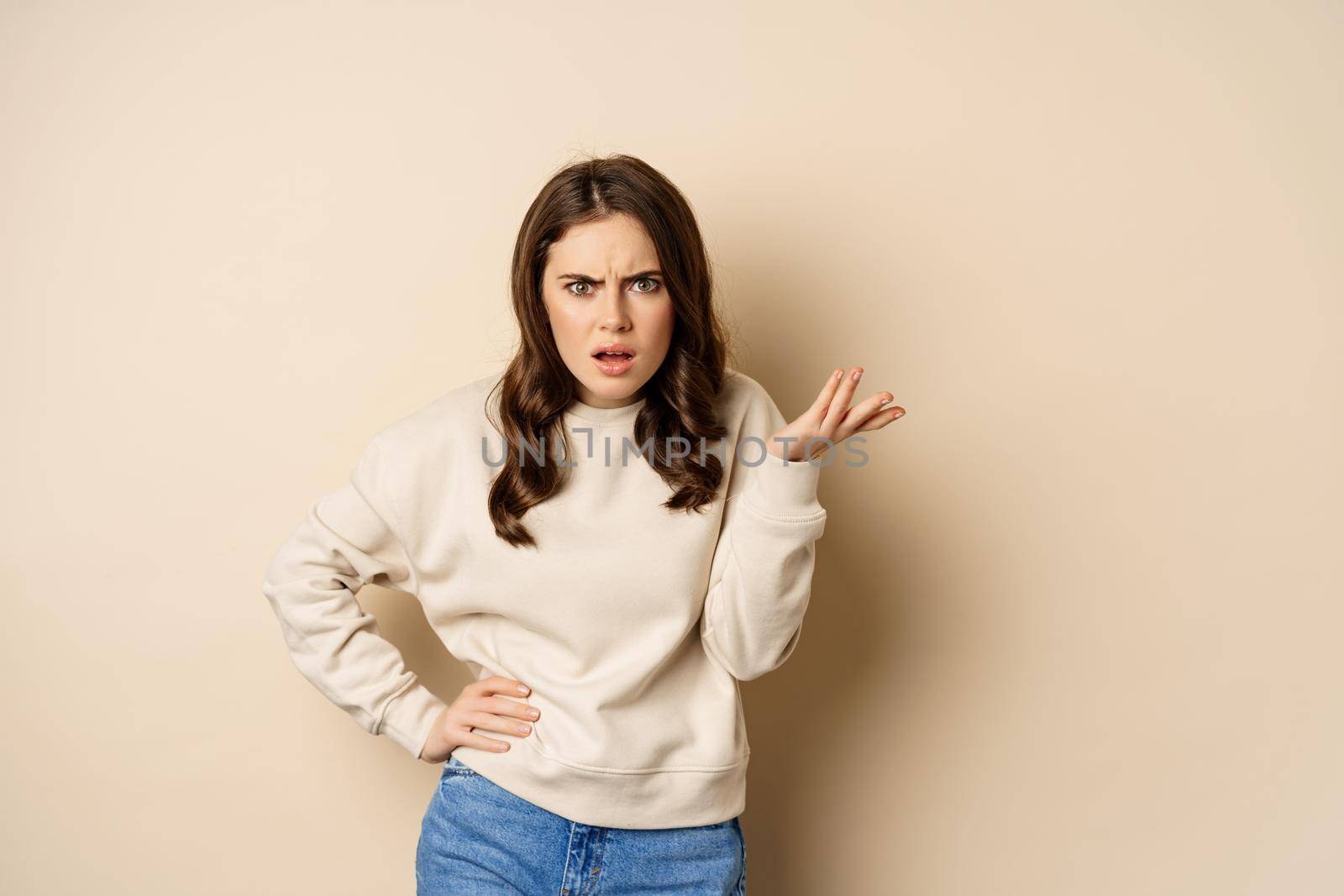 Frustrated and pissed-off woman, looking confused, arguing, standing over beige background.