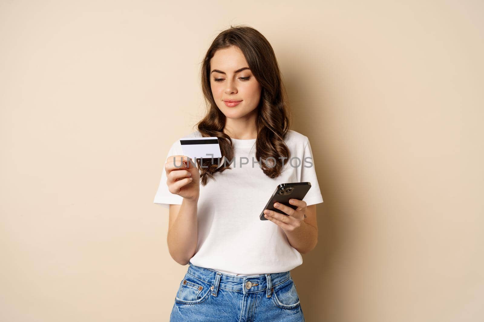 Image of young happy woman paying online, holding smartphone and credit card, enter info in application on mobile phone, standing against beige background.