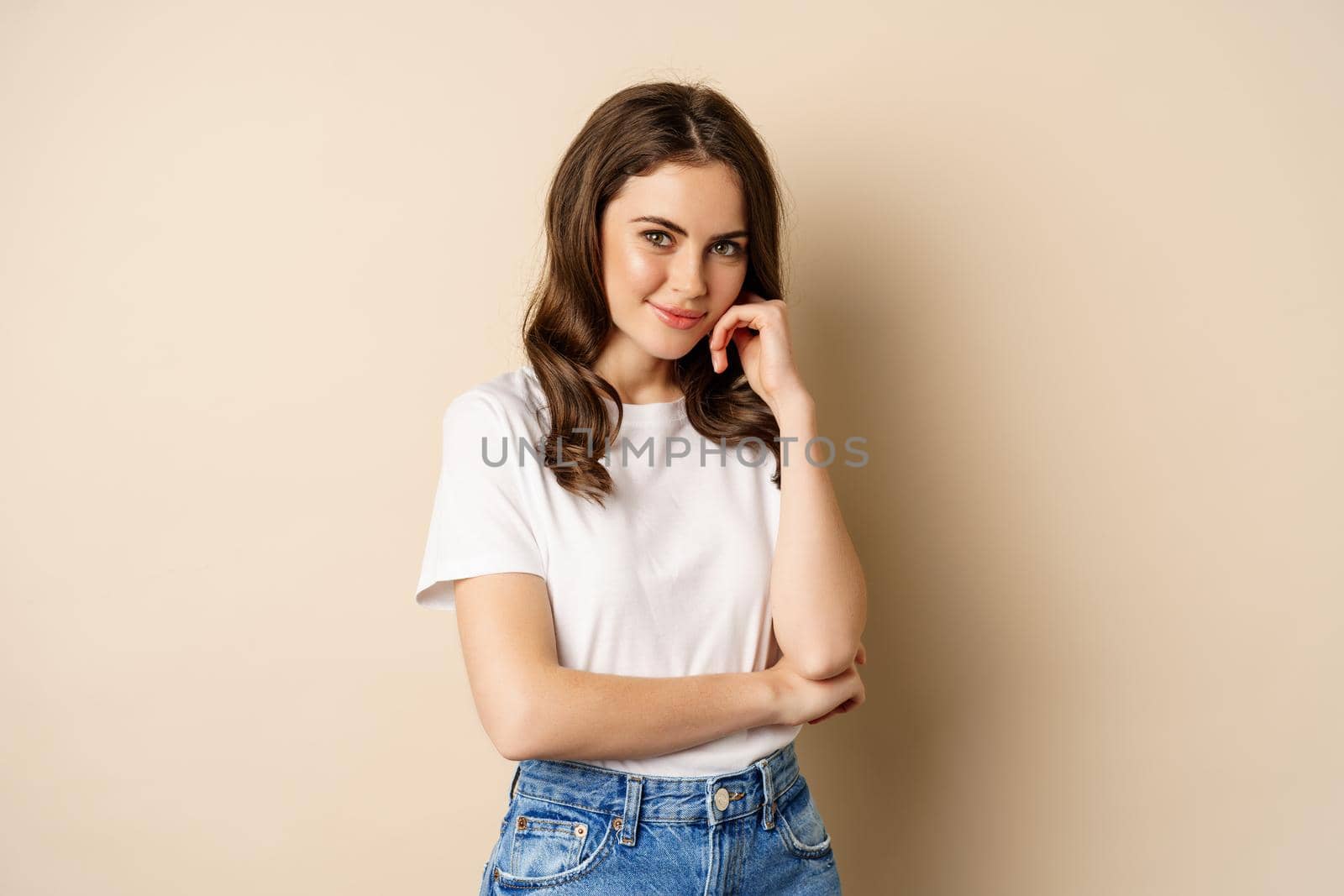 Portrait of beautiful young woman in white t-shirt, smiling and looking happy, posing against beige background.