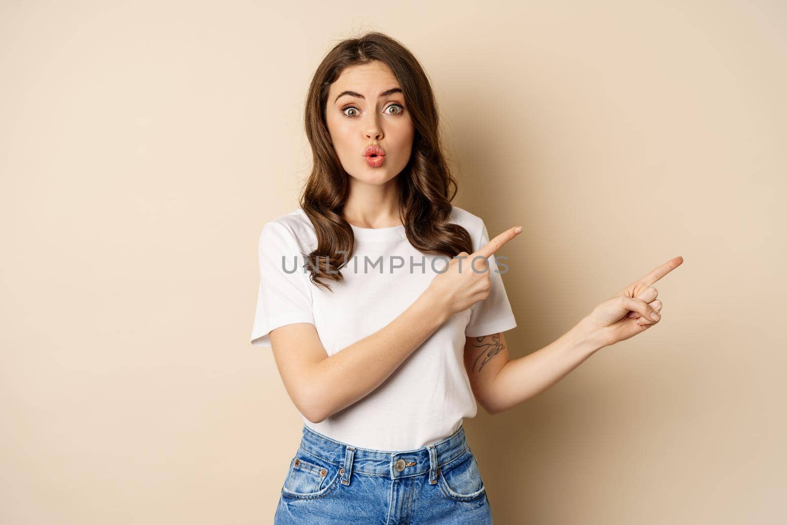 Excited girl pointing fingers right, showing promo with curious face expression, standing over beige background by Benzoix