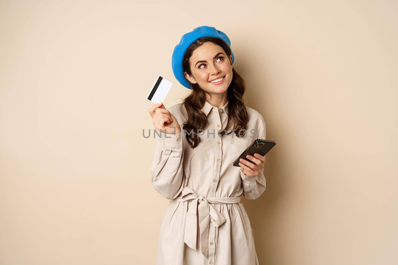 Portrait of trendy woman in coat holding smartphone, showing credit card, shopping online, order something on mobile phone, standing over beige background by Benzoix