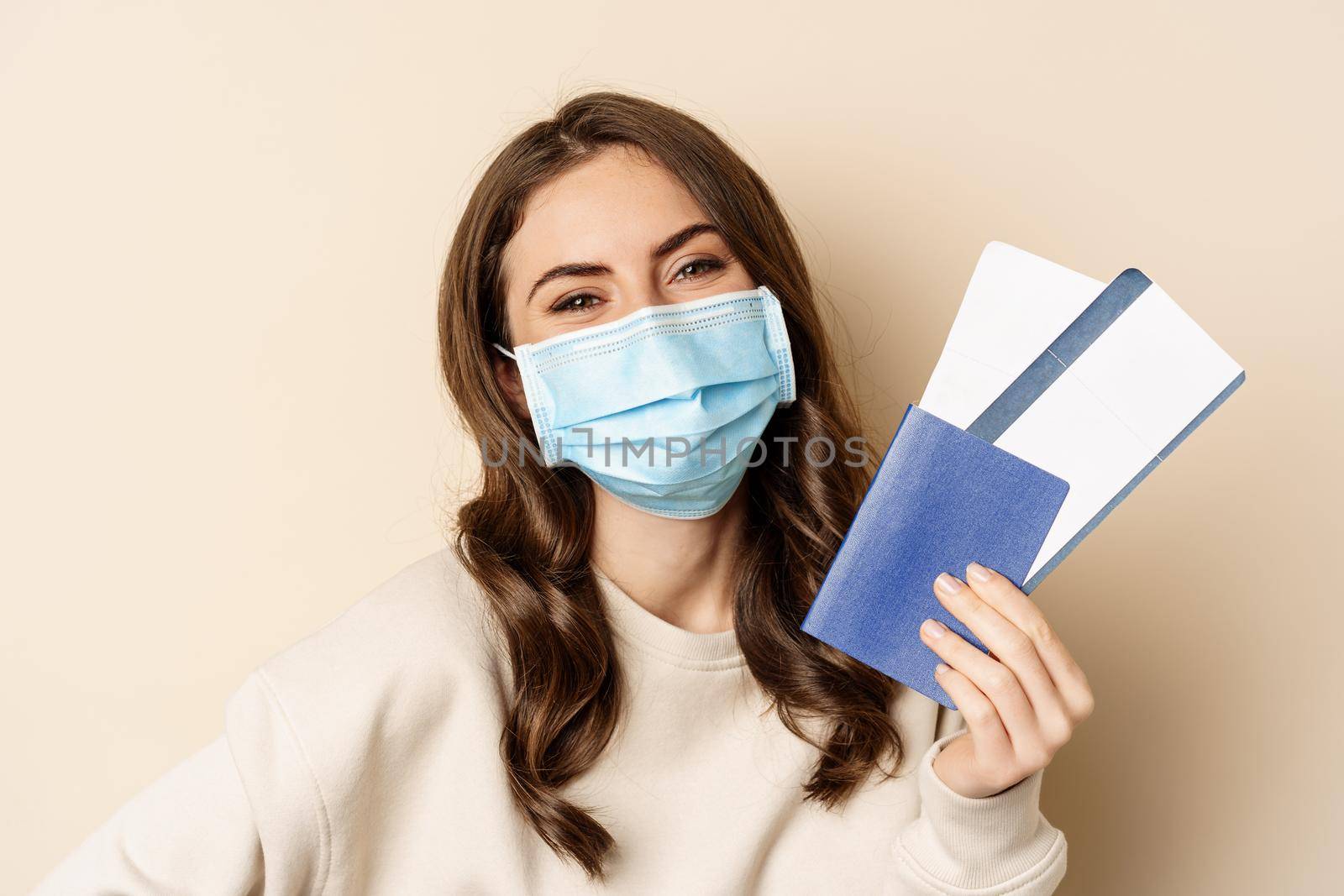 Travel and covid-19 pandemic. Close up portrait of smiling woman in medical mask, showing passport with two tickets, concept of tourism, beige background.