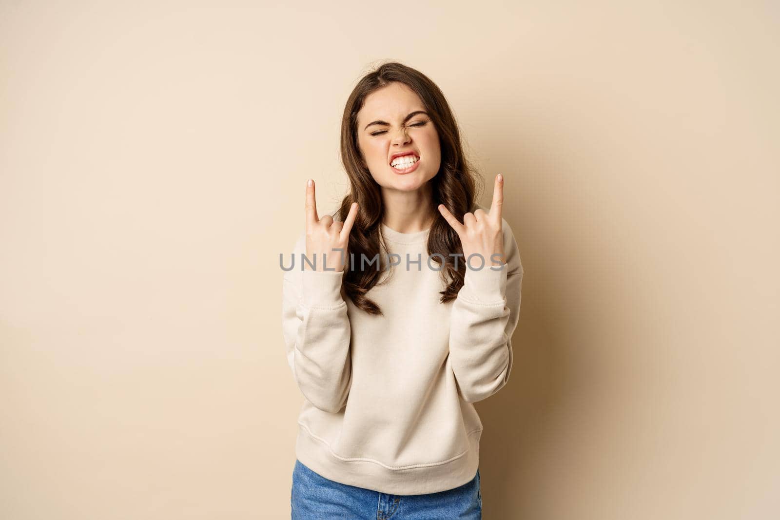 Happy young woman enjoying music, having fun, showing rock on, heavy metal finger horns gesture, standing over beige background.