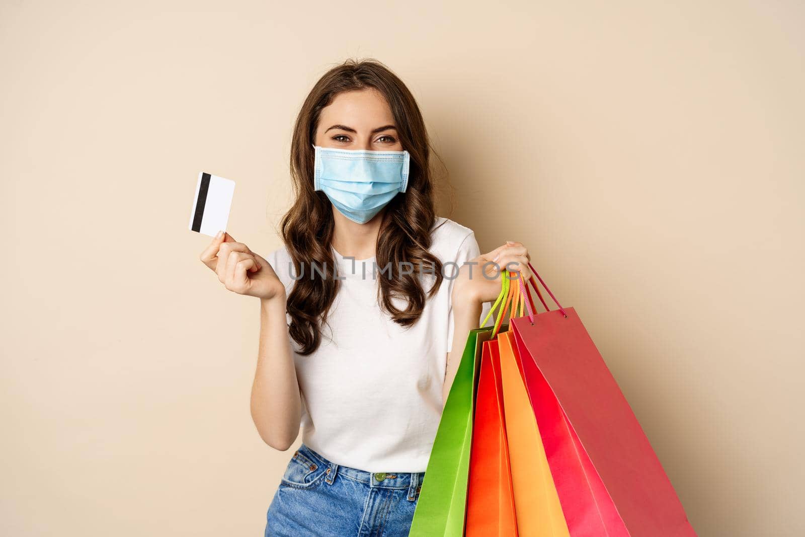 Covid-19 pandemic and lifestyle concept. Young woman posing in medical face mask with shopping bags from mall, holding credt card in hand, beige background.