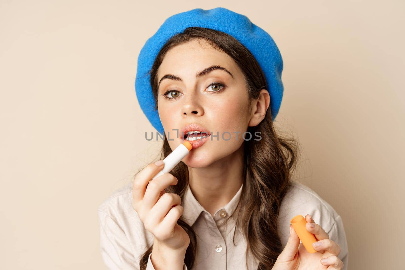 Close up portrait of young beatufiul woman looking in mirror and fixing her makeup, put on lipstick, standing over beige background. Cosmetics and women beauty concept