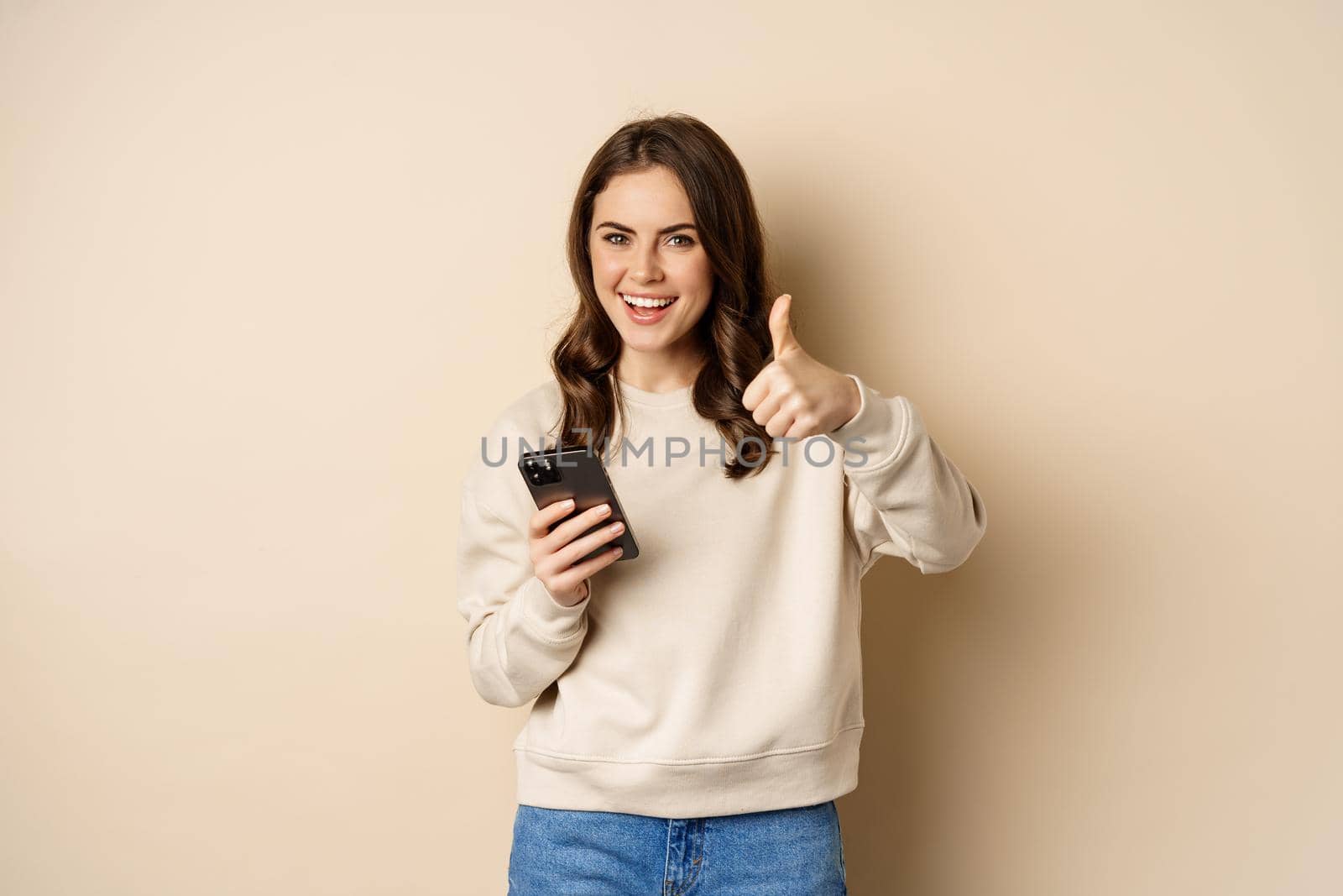 Young woman smiling, showing thumbs up while using mobile phone, smartphone app, standing over beige background by Benzoix