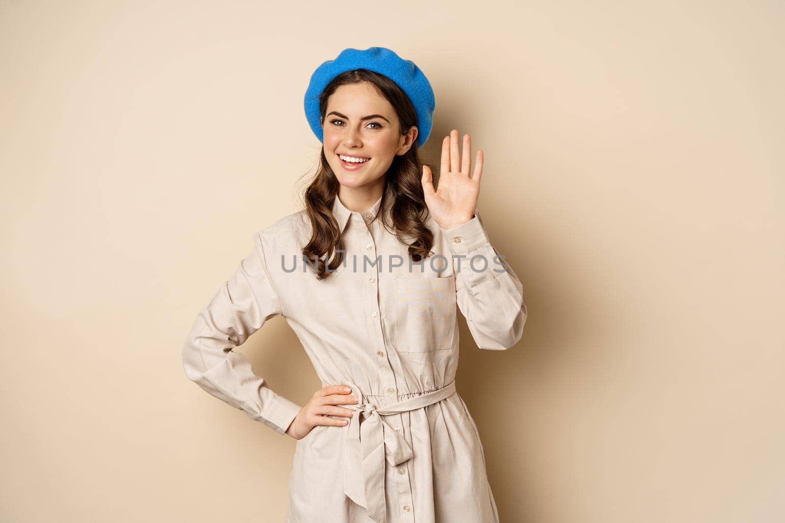 Image of beautiful friendly woman in outerwear, saying hello, waving hand and smiling, greeting you, standing over beige background.