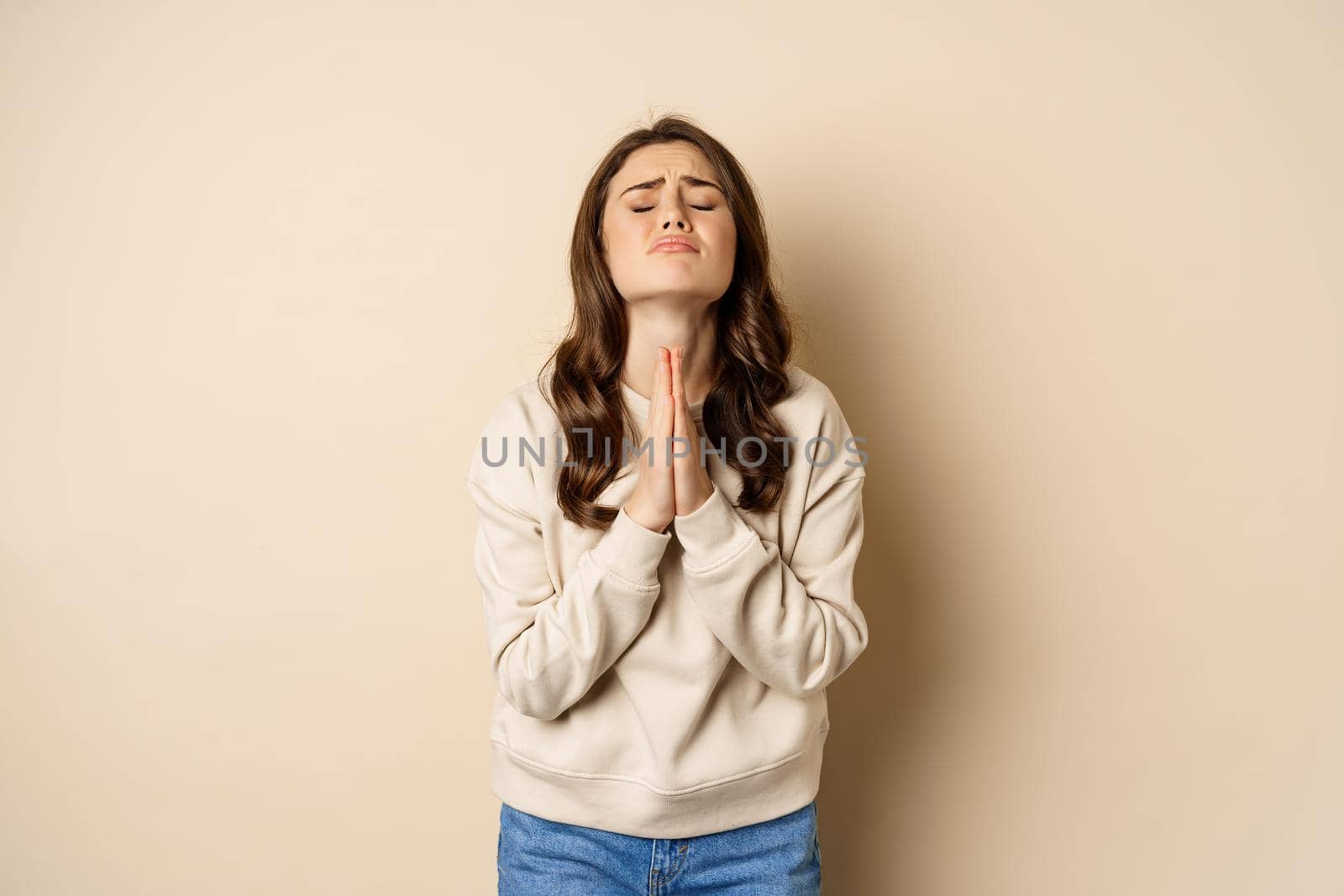 Woman begging and pleading god, supplicating, say please, standing desperate over beige background. Copy space