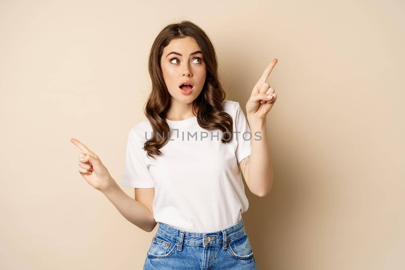 Happy authentic girl smiling, pointing fingers sideways, showing left and right banner, demonstrating promo, standing against beige background by Benzoix