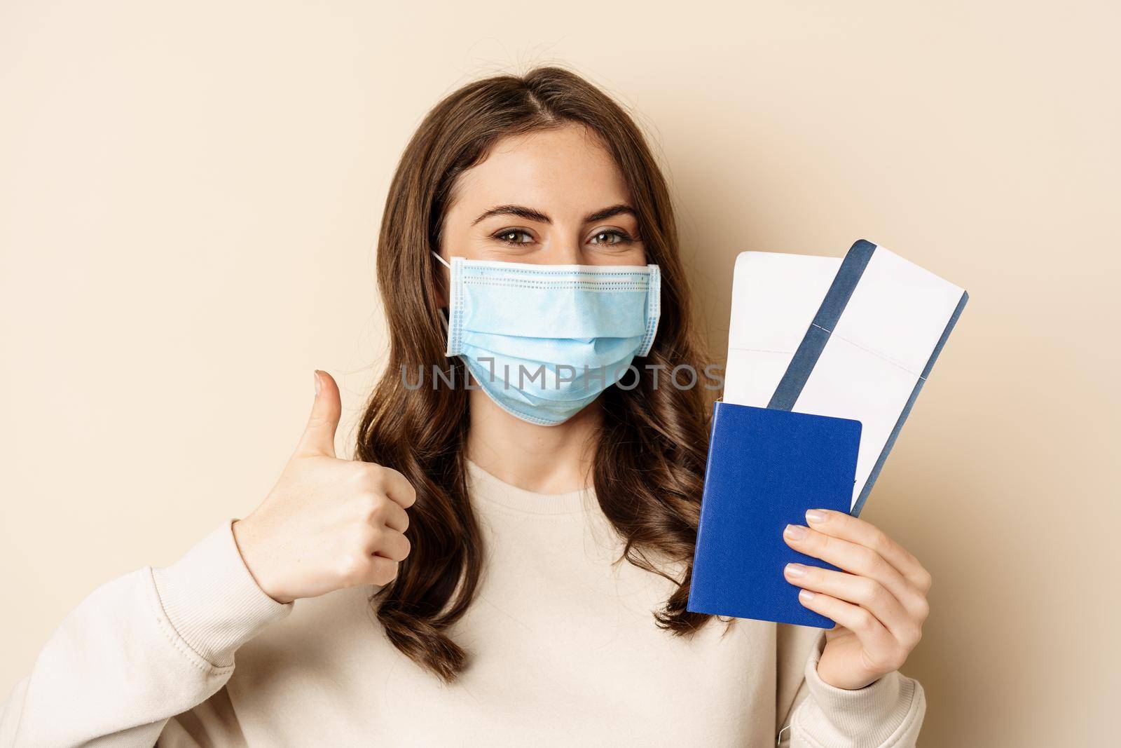 Travel and covid-19 pandemic. Happy woman tourist in medical mask, showing thumbs up and passport with two tickets, beige background.