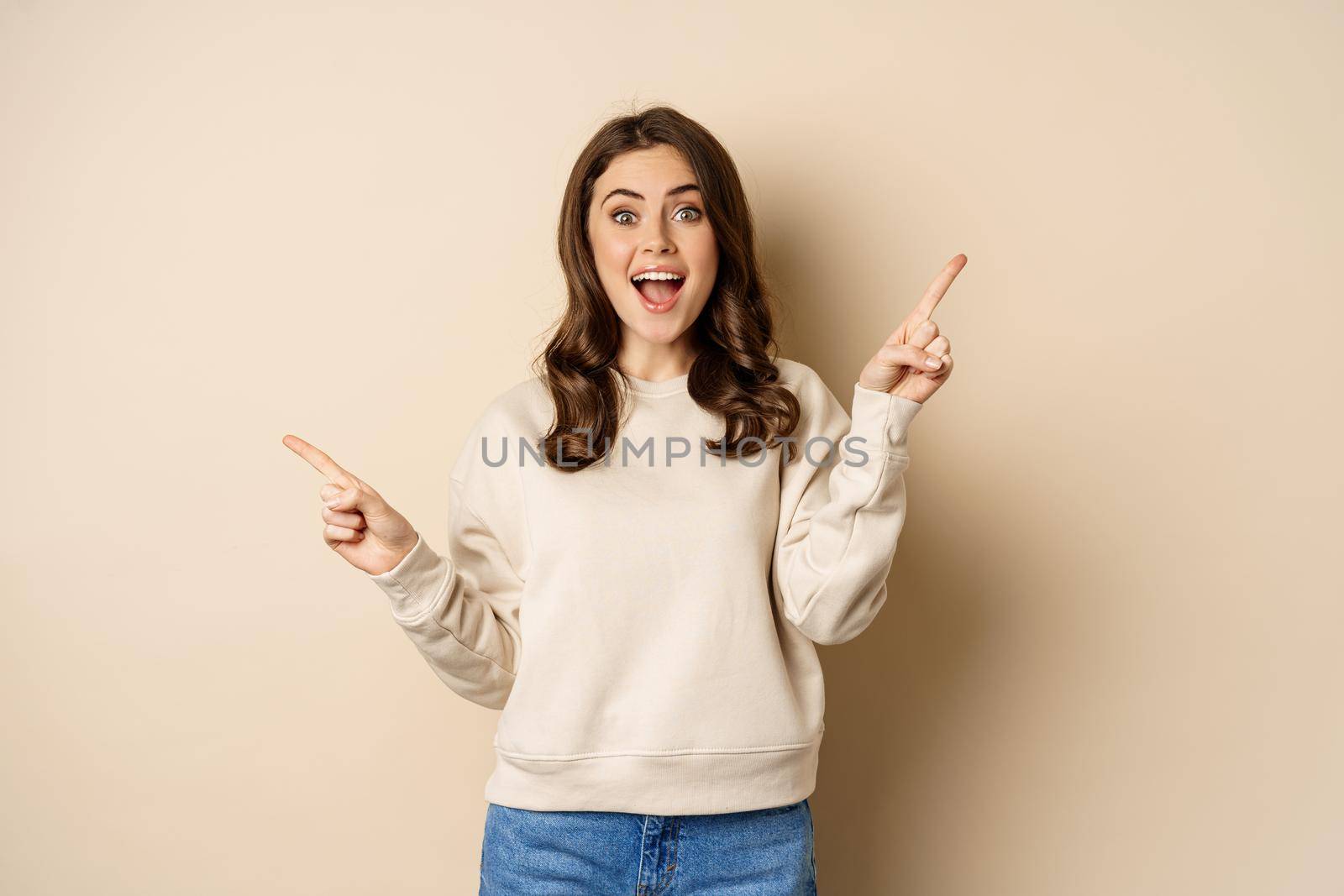 Cheerful caucasian woman showing directions, two ways, pointing sideways at variants, choices for customer, standing over beige background.