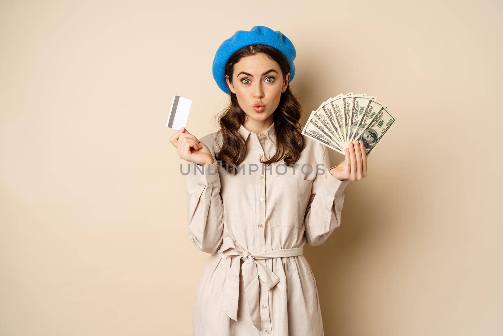 Microcredit and money concept. Young stylish woman showing credit card and dollars cash, smiling happy and satisfied, standing over beige background.
