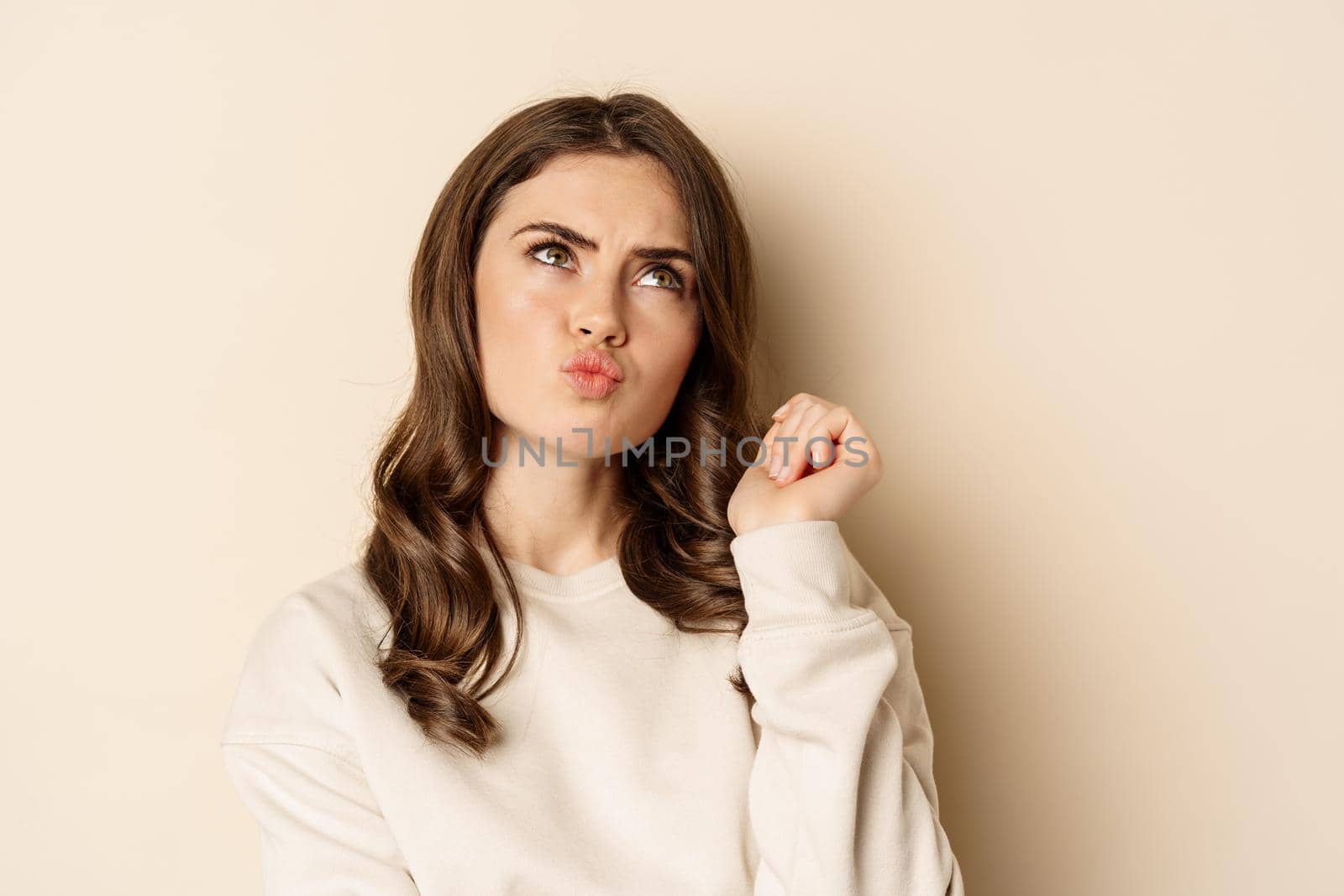 Thoughtful coquettish woman pucker lips and thinking, looking up complicated, standing over beige background, making decision.