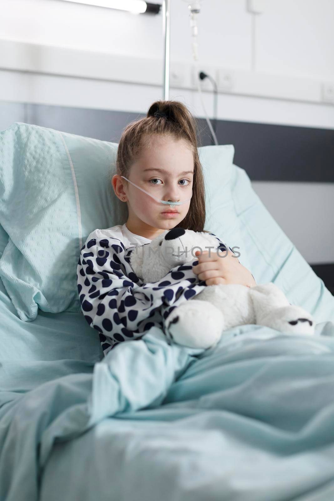 Unhealthy child in healthcare pediatric hospital patients treatment ward room while looking at camera. Sick kid holding teddybear and wearing oxygen tube while resting alone in pediatric clinic bed.