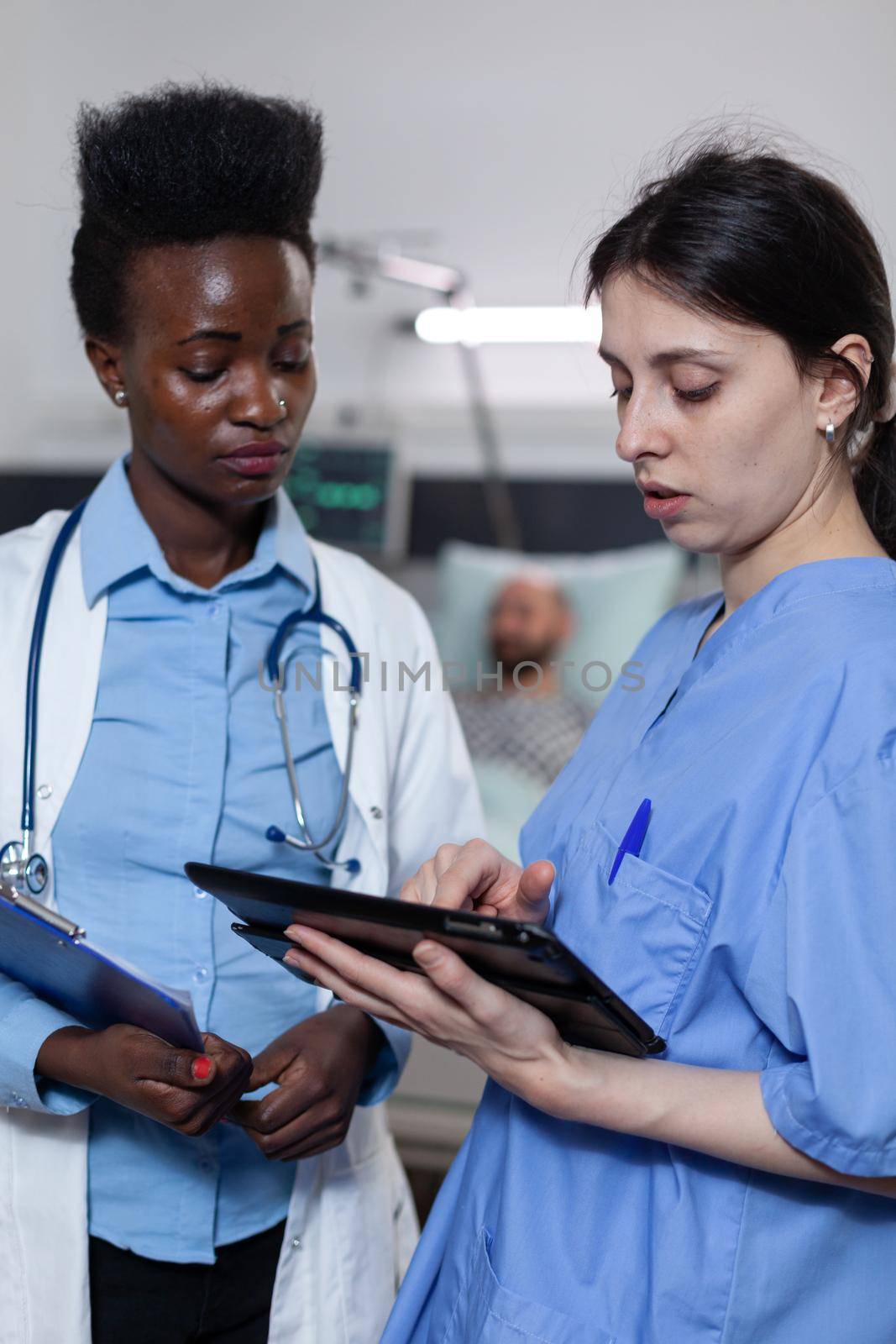 Nurse showing lab results from digital tablet to doctor holding clipboard with medical history in modern hospital ward. Health care specialists using modern technology for clinical diagnosis.