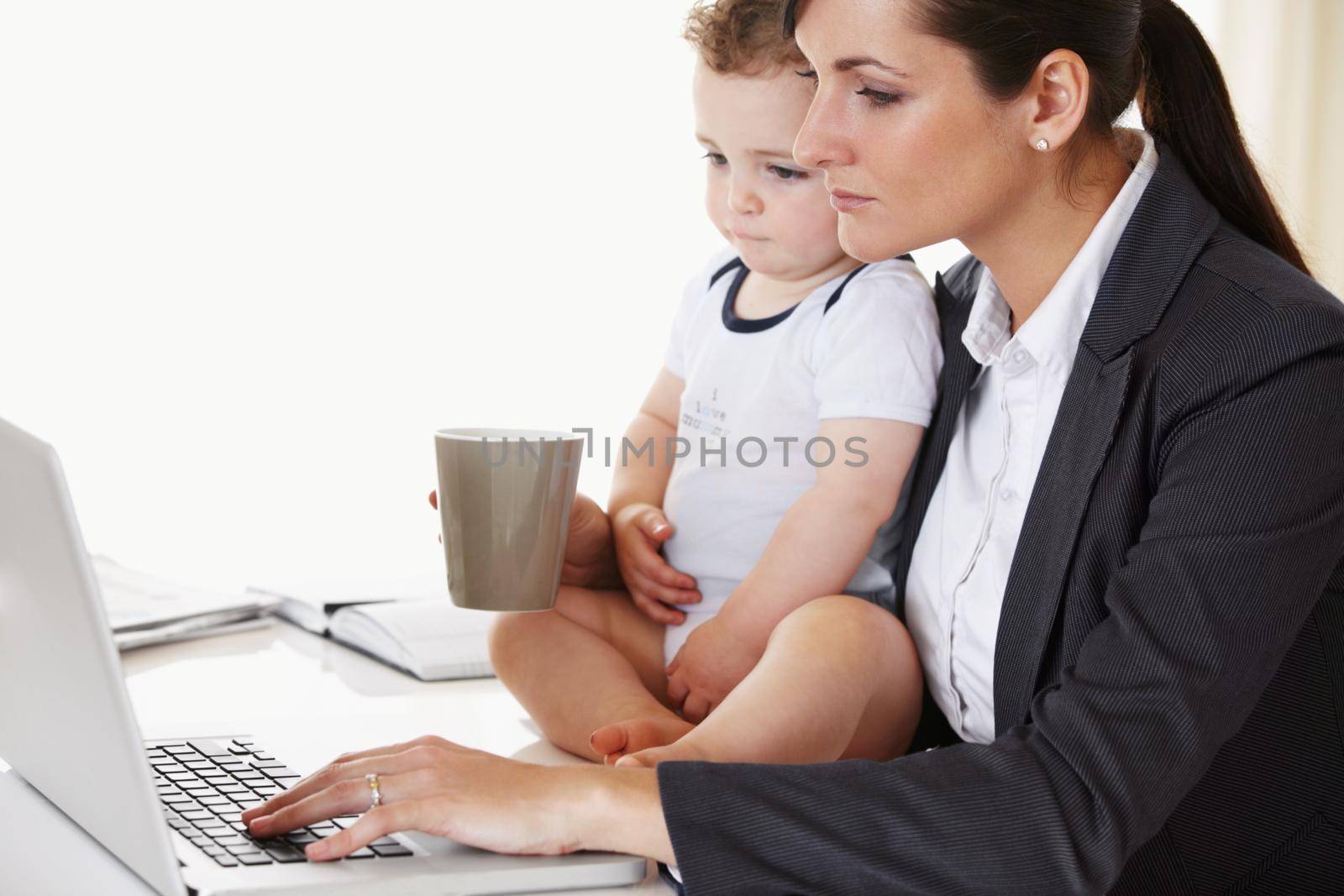 Young working mother holding a baby while working and drinking coffee.