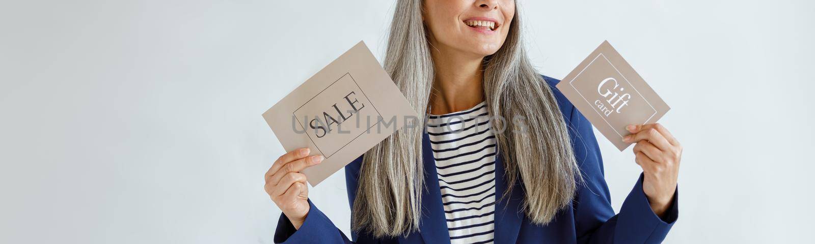 Cheerful middle aged Asian woman holds gift card and Sale sign standing in studio by Yaroslav_astakhov