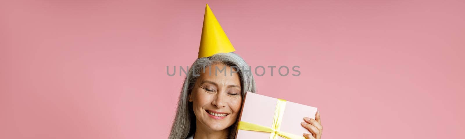 Happy mature Asian lady with long grey hair and yellow party hat holds gift box posing on pink background in studio