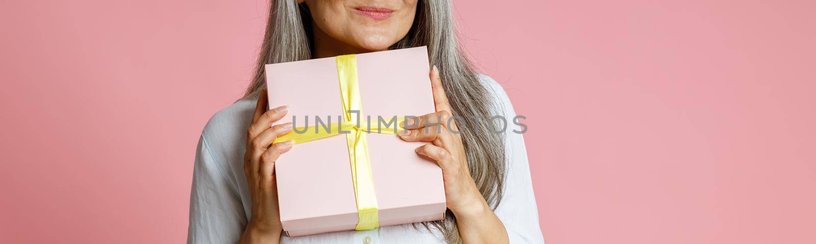 Positive mature female model with long silver hair holds gift box posing on pink background in studio closeup