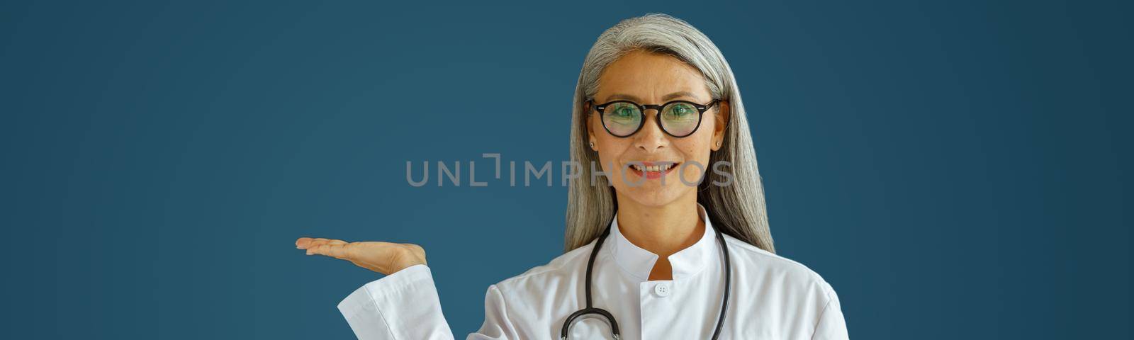 Positive middle aged lady doctor in white robe with glasses and stethoscope points aside standing on blue background in studio. Professional medical staff