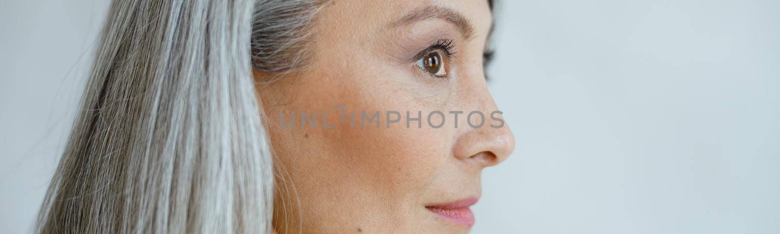 Middle aged Asian woman with beautiful straight hoary hair poses on light grey background in studio closeup side view. Mature beauty lifestyle