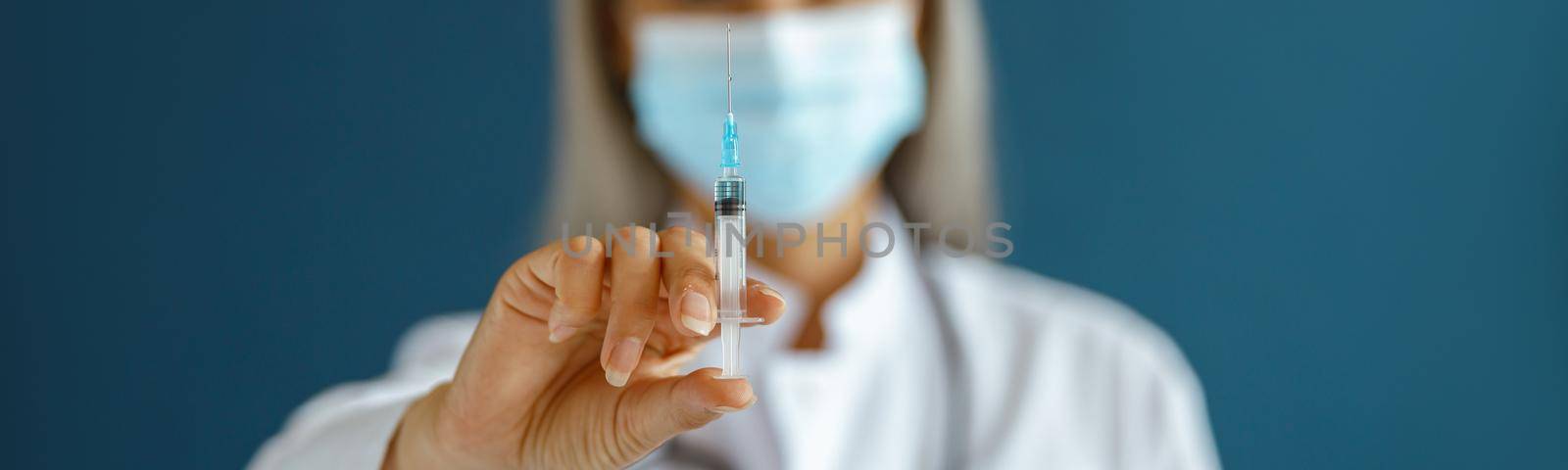 Grey haired Asian woman doctor in white coat with disposable mask stands on blue background in studio, focus on hand with syringe. Vaccination day