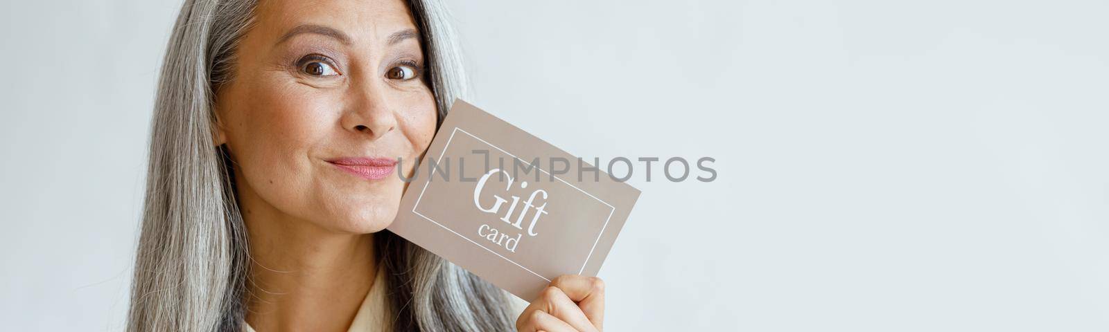 Joyful grey haired Asian woman holds gift card near face posing on light grey background in studio, space for text. Shopping certificate