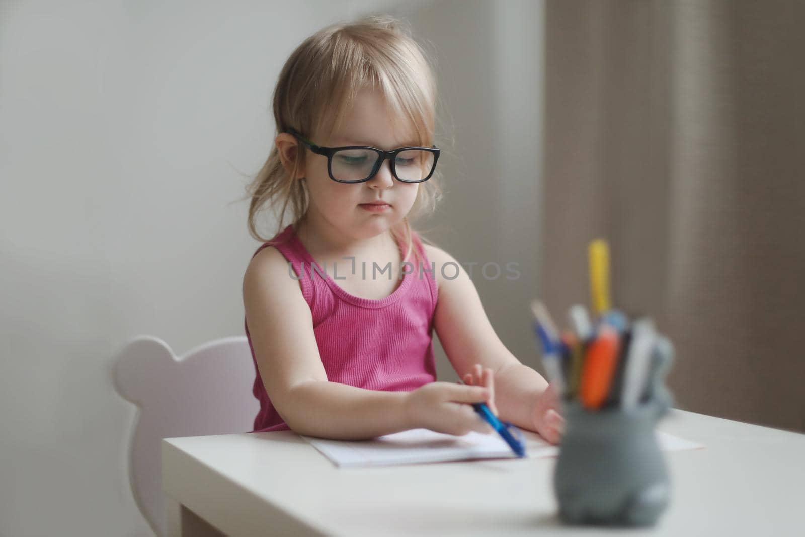 small funny girl in eyeglasses writing and drawing at the table.