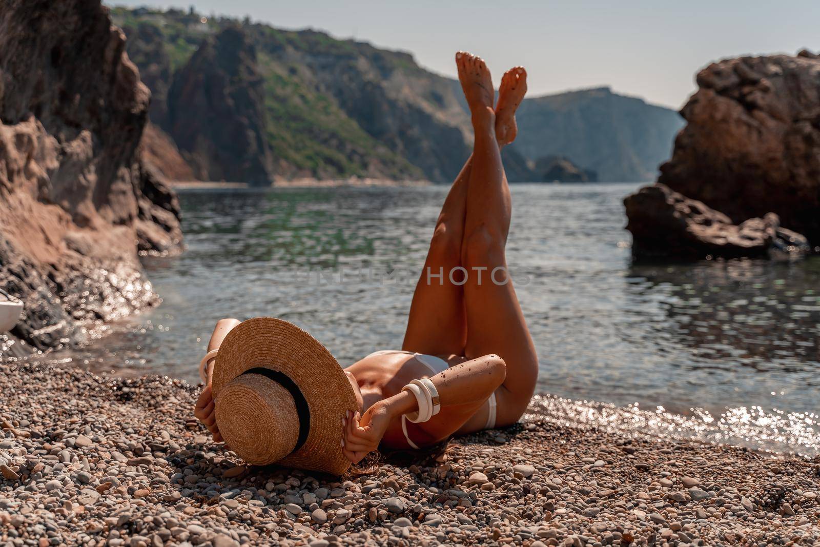 Girl in hat lying on beach with legs up. Fashionable young woman covering with straw hat, relaxing on sandy beach near sea. Summer vacation and travel. Mindfulness and carefree.
