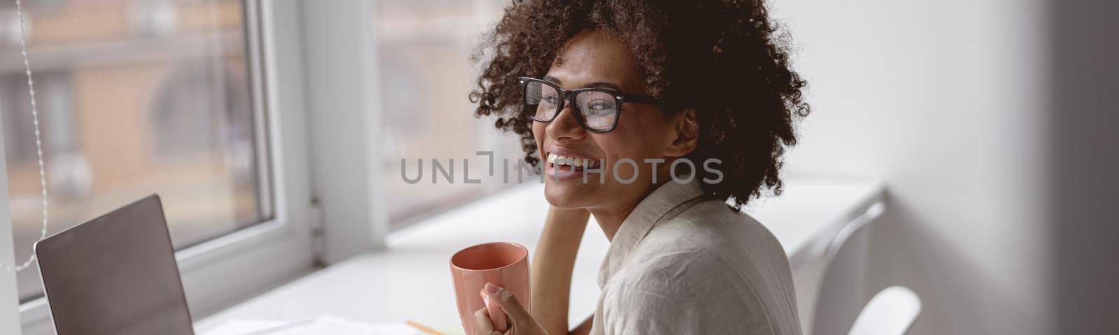 Smiling multiethnic woman wearing glasses and sitting at the desk with laptop while enjoying coffee