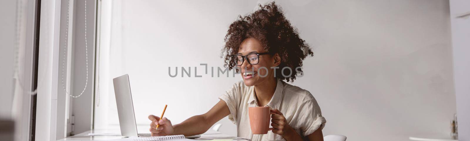 Happy African American lady sitting near the window and working with a laptop while writing in a notebook
