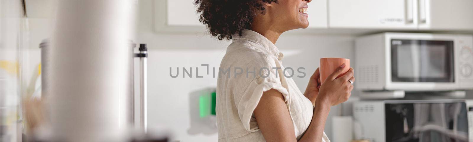Side view of smiling multiethnic woman holding cup of coffee while standing at office kitchen