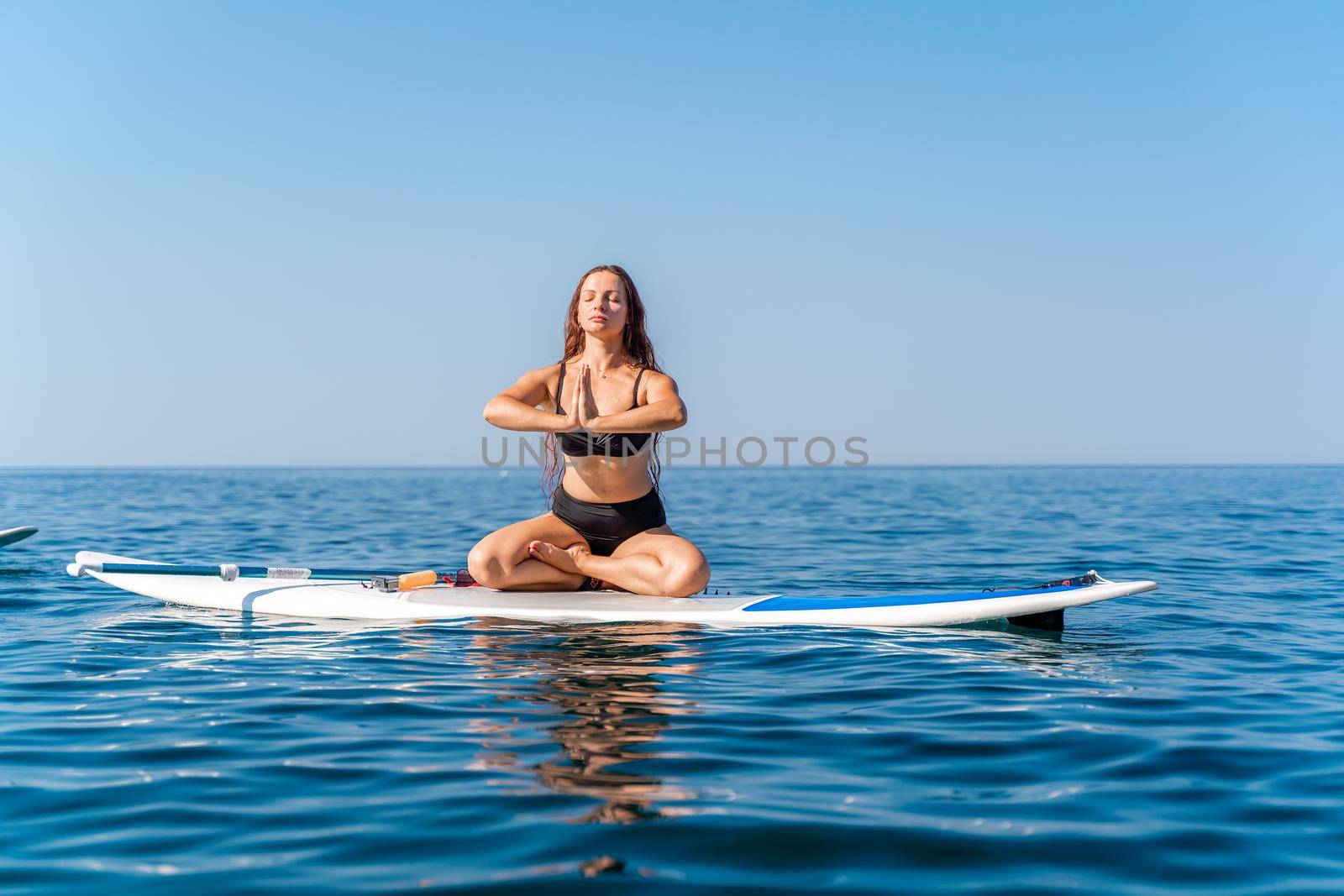 Sporty girl on a glanders surfboard in the sea on a sunny summer day. In a striped swimsuit, he sits in the lotus position with his eyes closed
