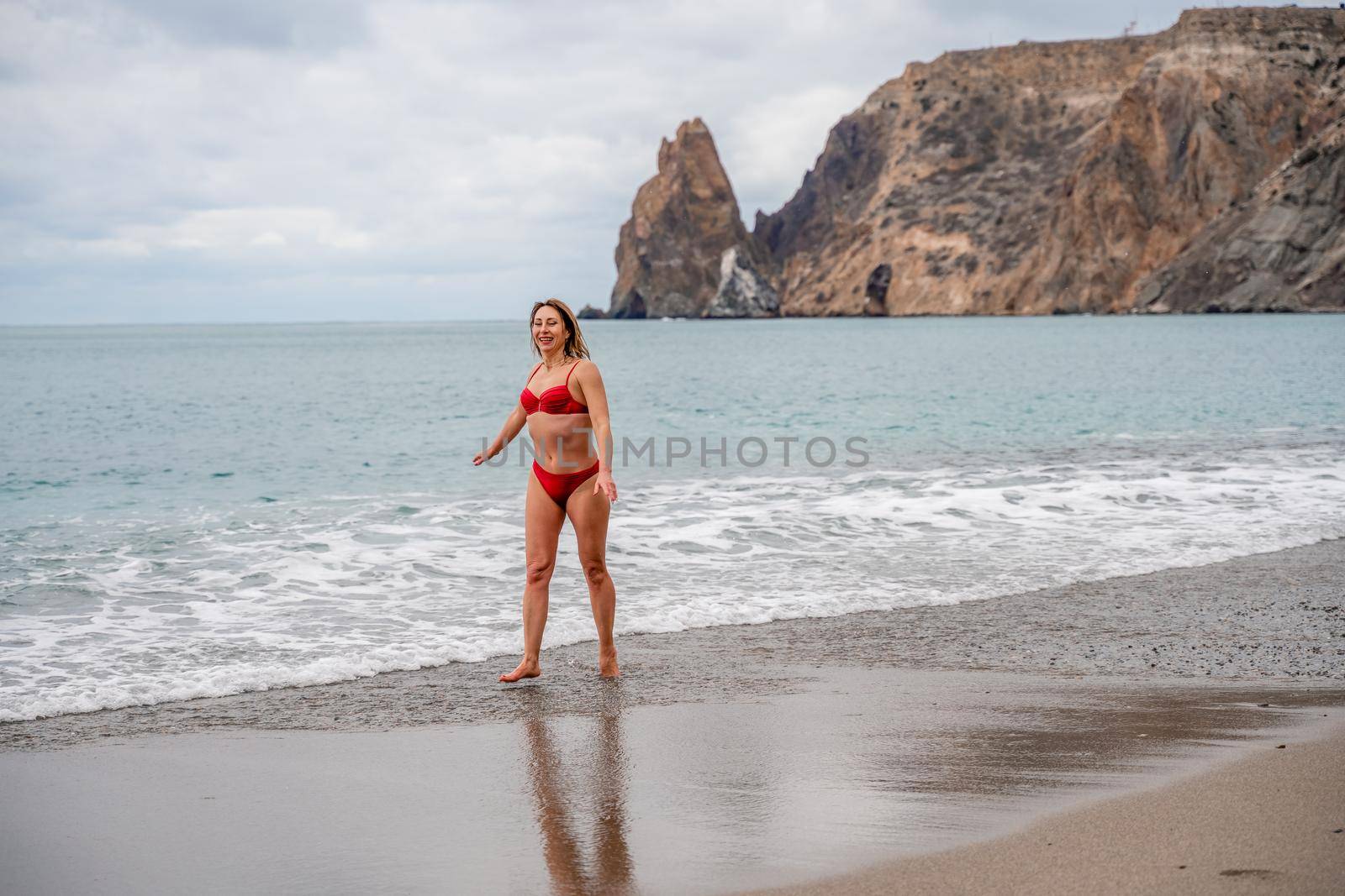 A middle-aged woman with a good figure in a red swimsuit on a pebble beach, running along the shore in the foam of the waves.