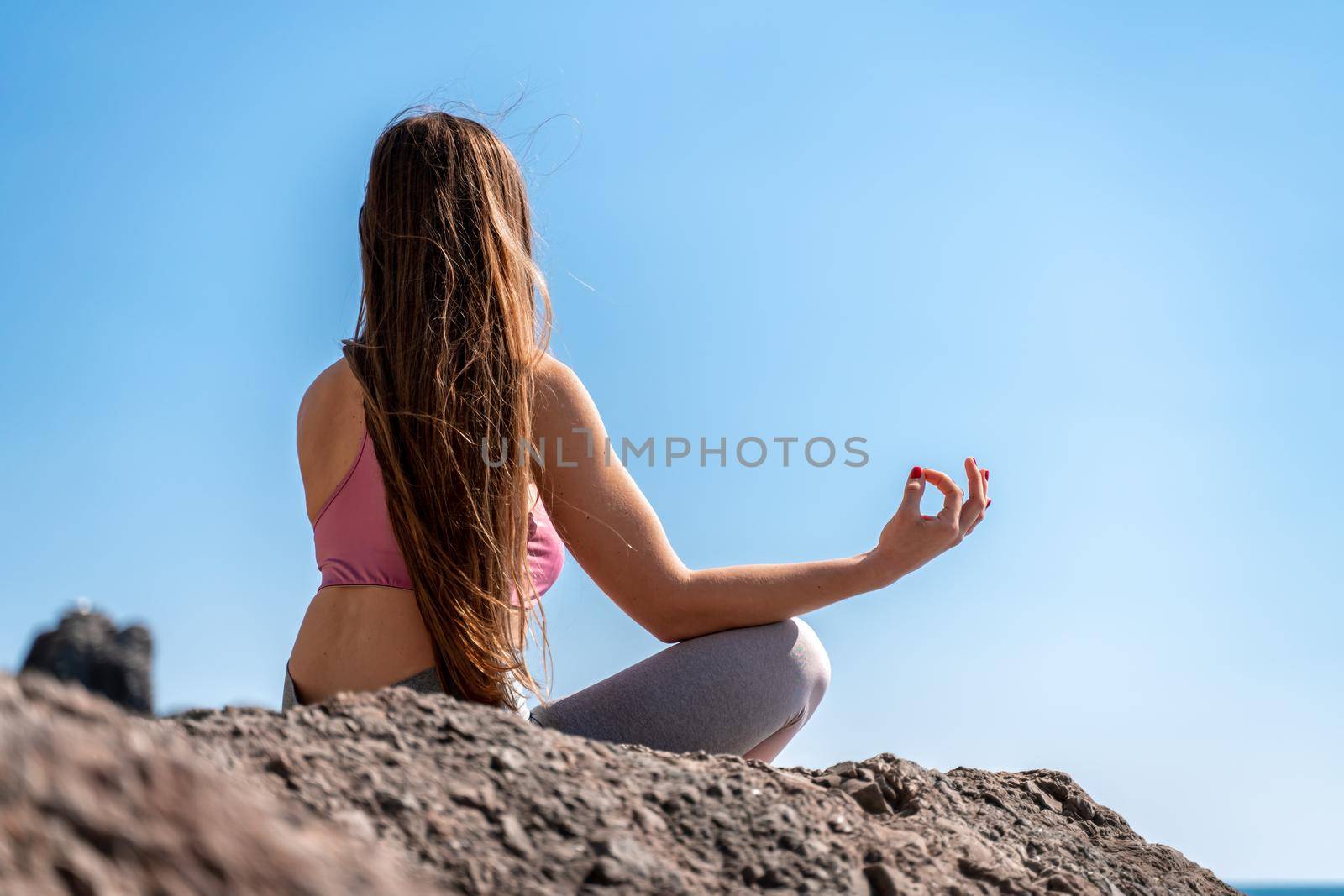A young woman in gray leggings and a pink top with long loose hair does yoga outdoors by the sea on a sunny day. Women's yoga, fitness, Pilates. The concept of a healthy lifestyle, harmony. by Matiunina