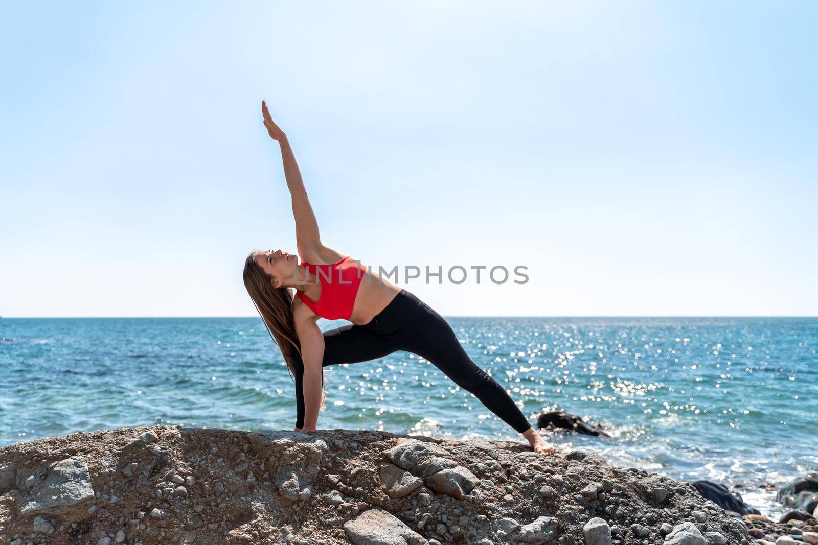 Young woman in black leggings and a red top with long flowing hair practicing stretching outdoors by the sea on a sunny day. Women's yoga, fitness, pilates. The concept of a healthy lifestyle, harmony