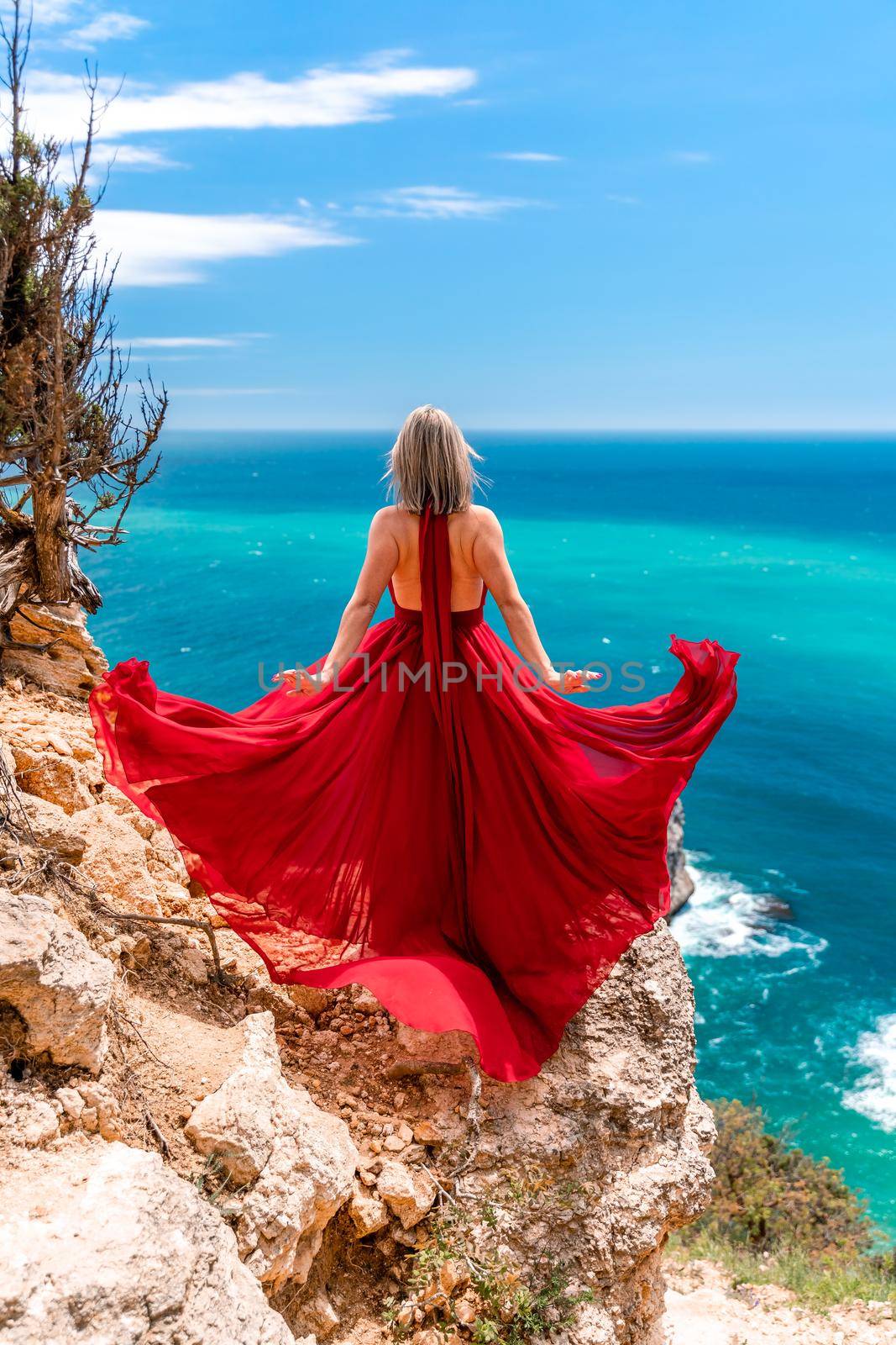 A girl with loose hair in a long red dress descends the stairs between the yellow rocks overlooking the sea. A rock can be seen in the sea. Sunny path on the sea from the rising sun by Matiunina