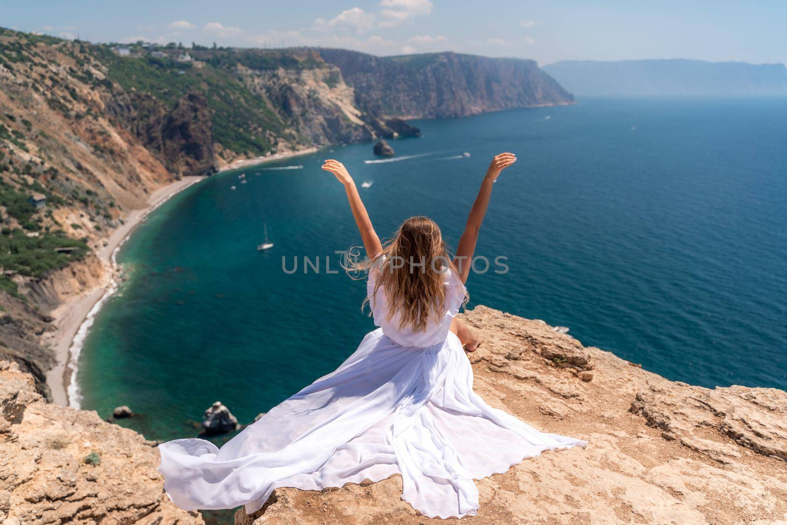 Portrait of a young woman on the beach by the sea, sitting with raised hands in a white dress and hat. Back photo by Matiunina