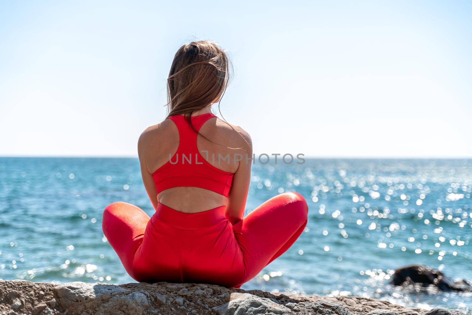A young woman in red leggings and a red top with long loose hair practices yoga outdoors by the sea on a sunny day. Women's yoga, fitness, Pilates. The concept of a healthy lifestyle, harmony