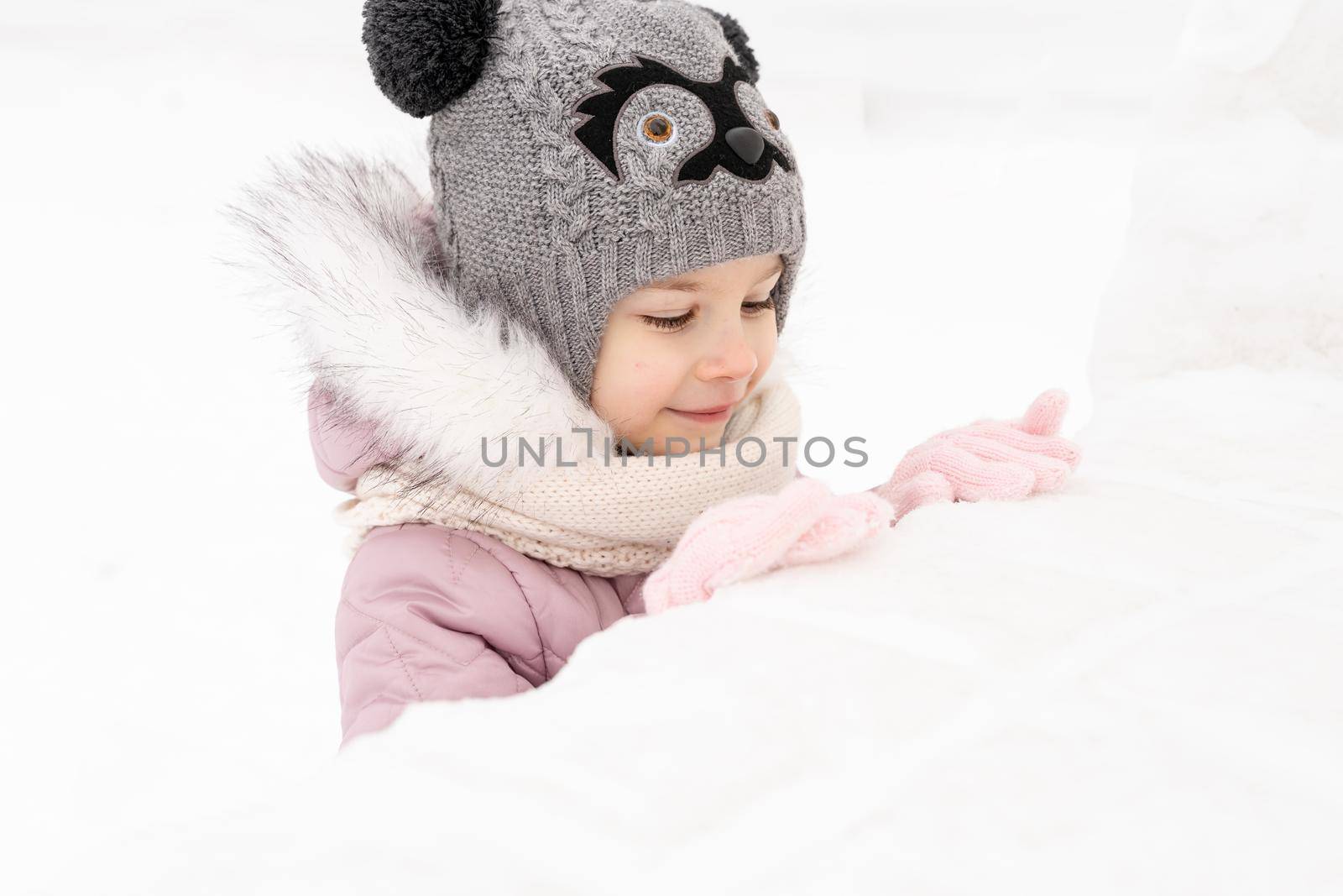 little girl in a knitted hat and scarf and a snow fort by Lena_Ogurtsova