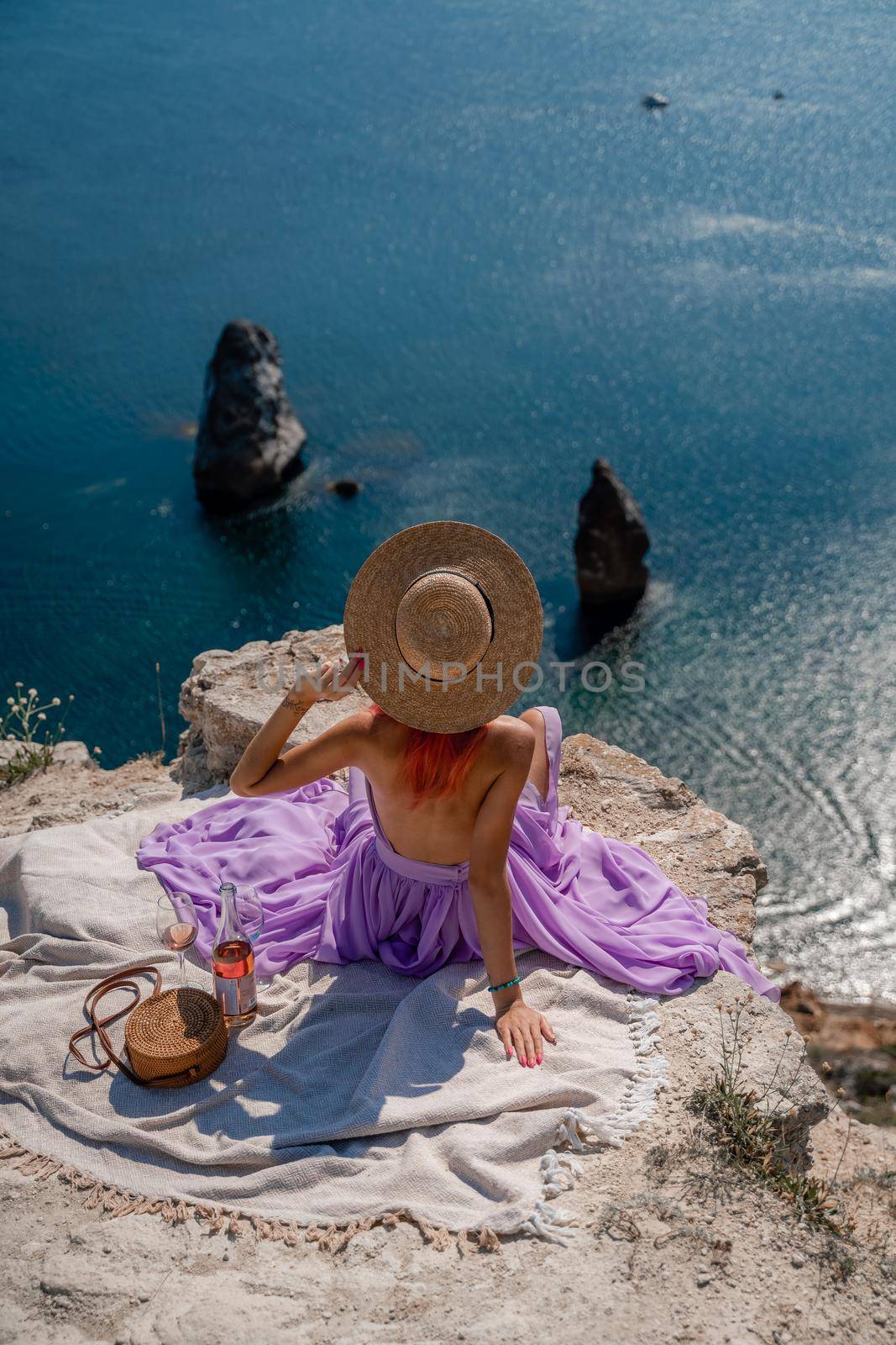 A girl in a hat sits with her back to the viewer on a picnic blanket in a pink dress and holds a hat with her hands. Champagne, two glasses, Summit on the mountain against the background of the sea and rocks in the sea.