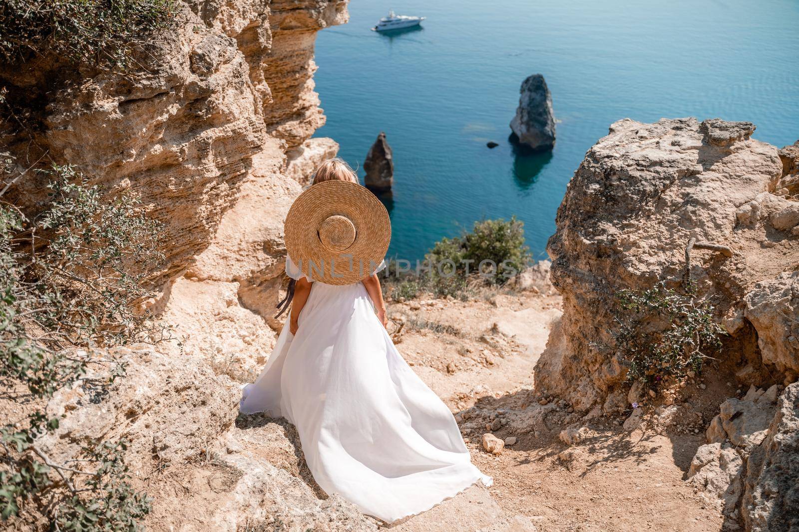A girl with loose hair in a long white dress descends the stairs between the yellow rocks overlooking the sea. A rock can be seen in the sea. Sunny path on the sea from the rising sun by Matiunina