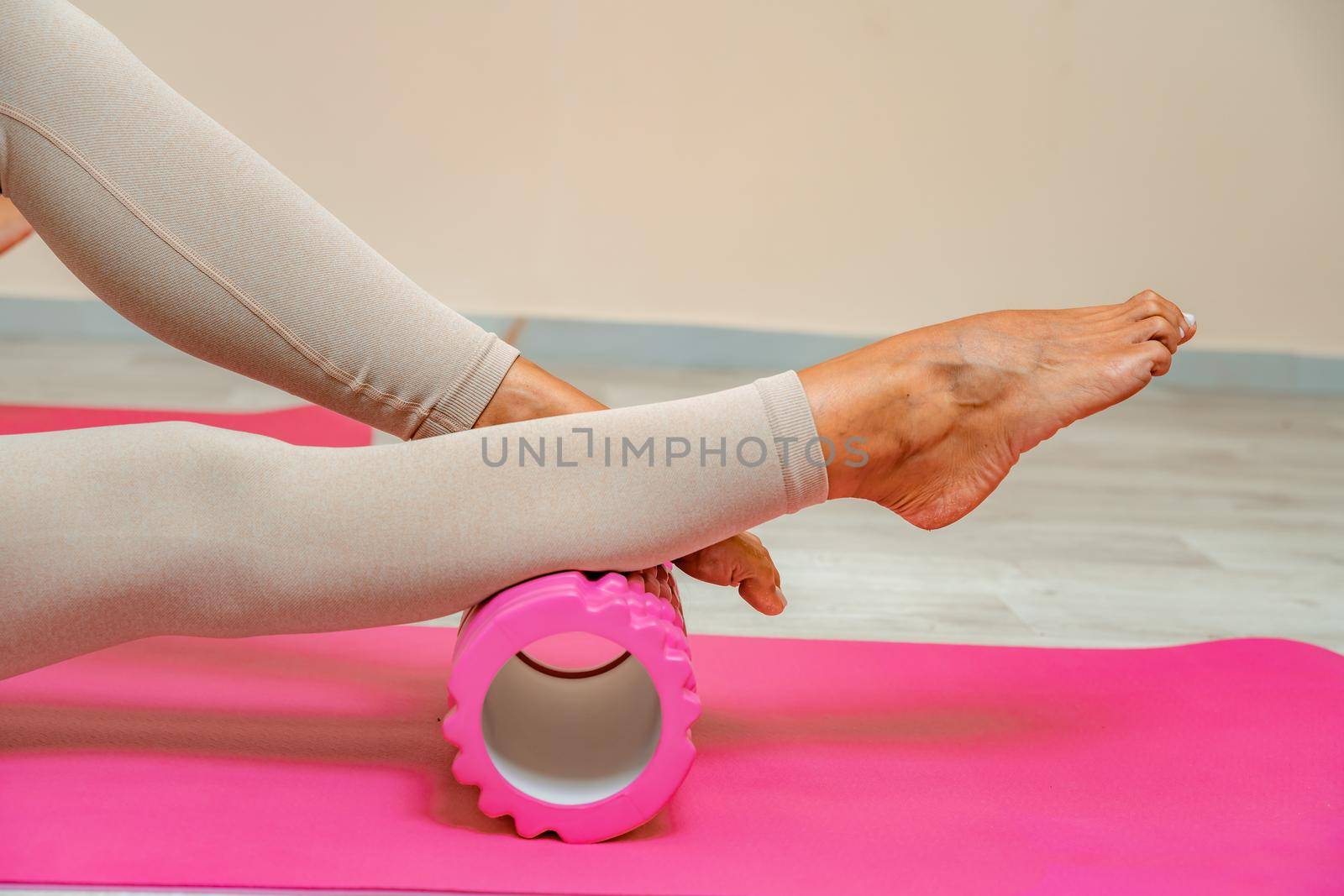 Fit athletic women in beige sportswear ride on a pink foam myofascial massage roller, massaging their muscles on the floor of a yoga mat in the gym. Active girls do a fitness workout in the gym. MFR by Matiunina