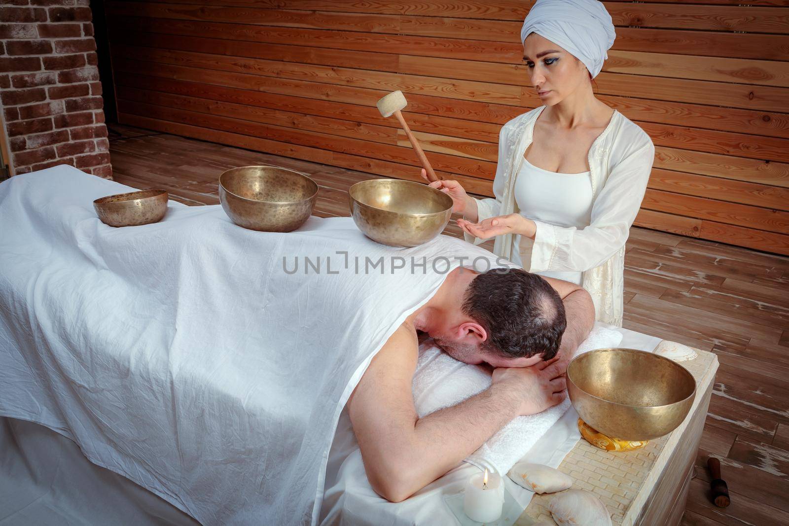 A woman performs Tibetan singing bowl therapy with a man lying under a white sheet. Relaxing meditation.