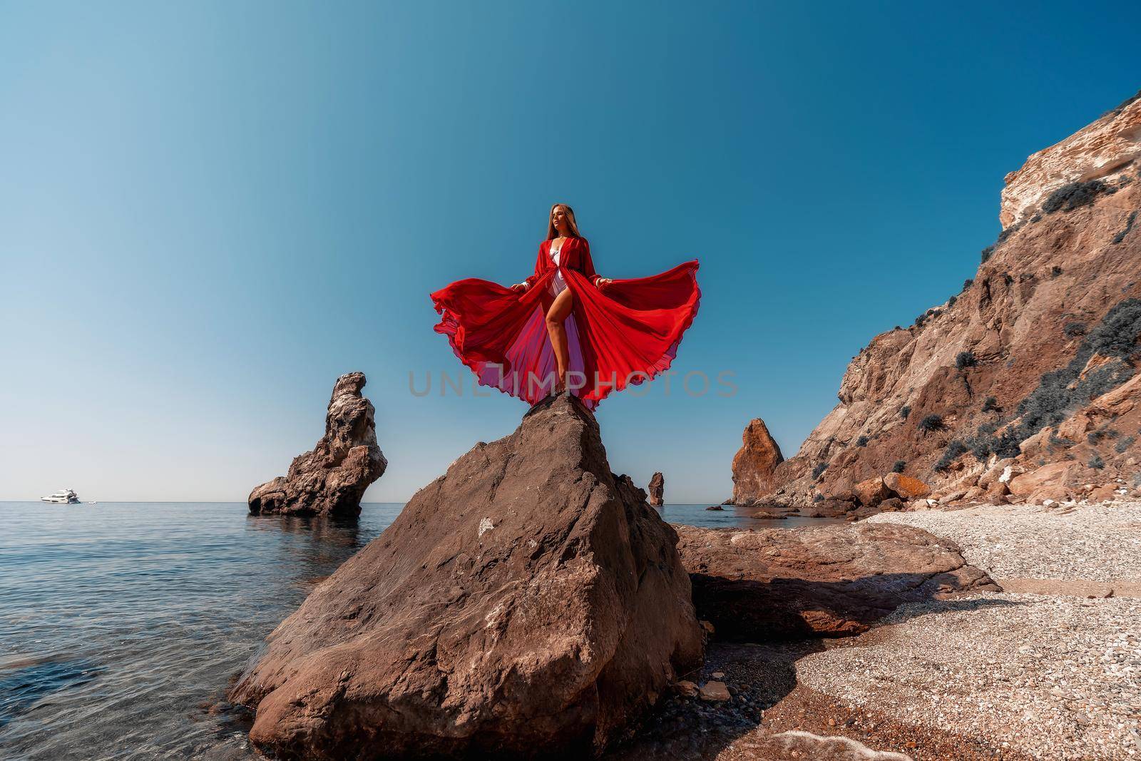 Beautiful woman posing in a luxurious red dress for the beach and in a white swimsuit with a long train, standing on a cliff against the sea. Around the cliff, the seashore