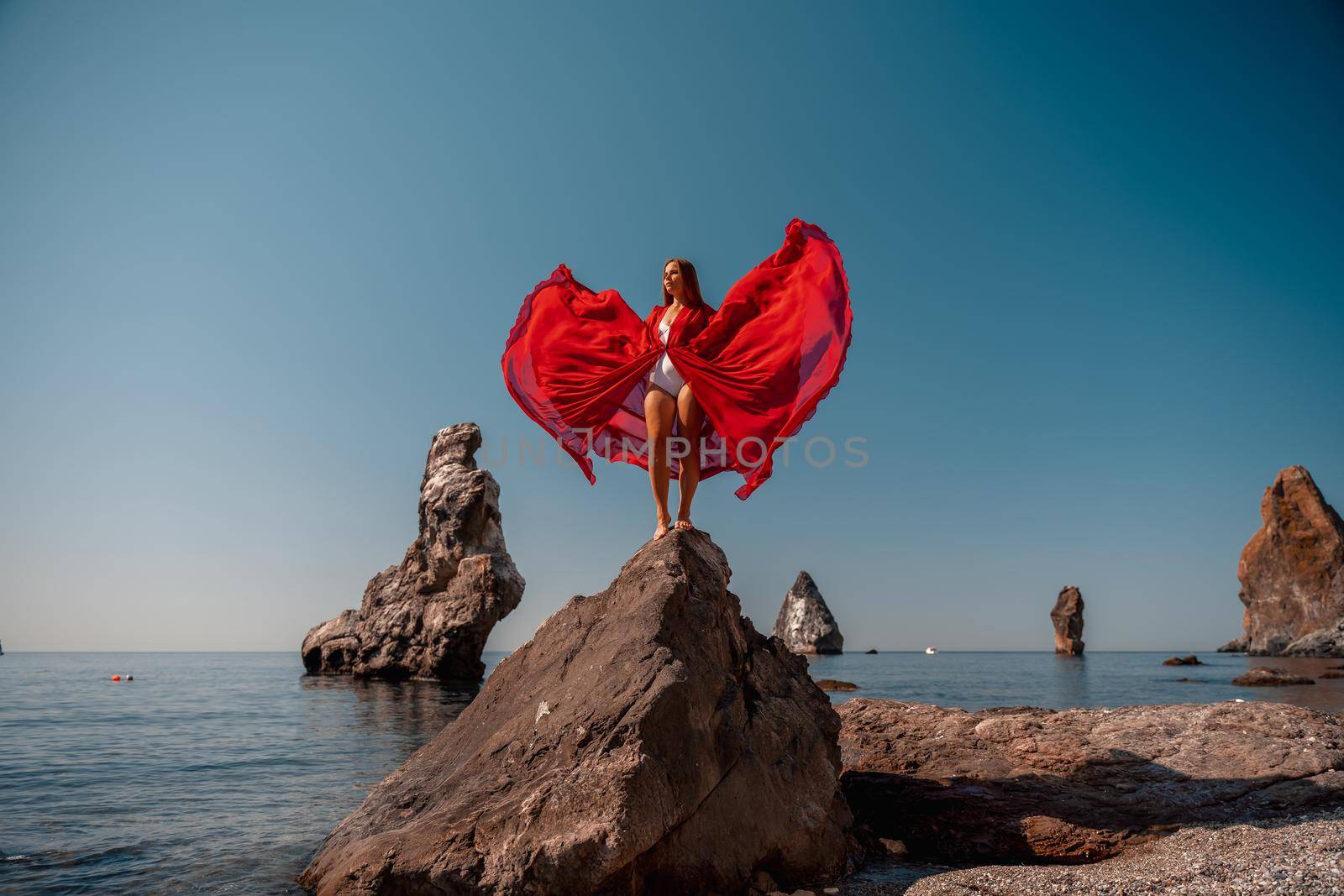 Beautiful woman posing in a luxurious red dress for the beach and in a white swimsuit with a long train, standing on a cliff against the sea. Around the cliff, the seashore