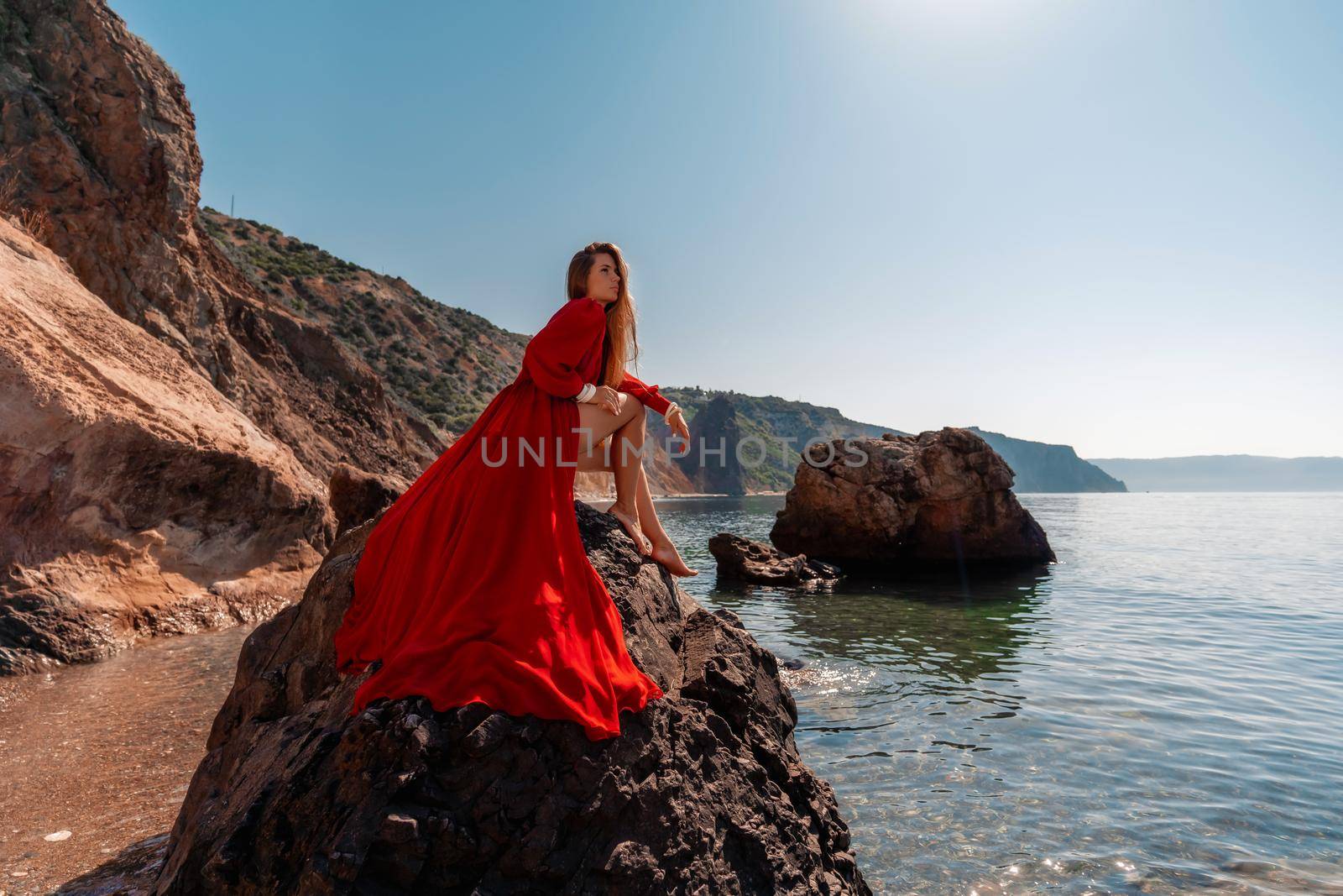 Beautiful sensual woman in a flying red dress and long hair, sitting on a rock above the beautiful sea in a large bay