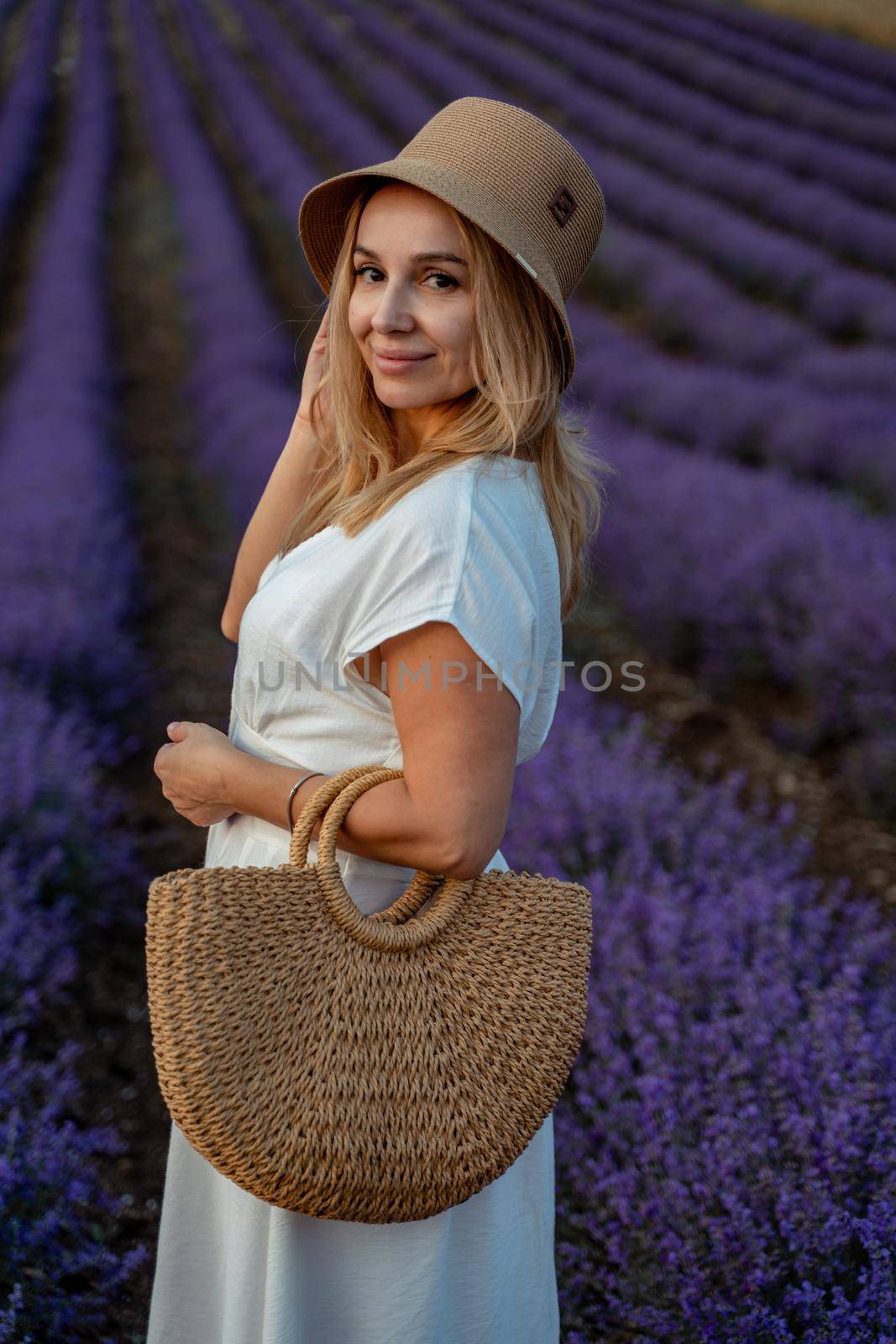 A middle-aged woman, blonde in a white dress and hat, walks through a lavender field with a basket.