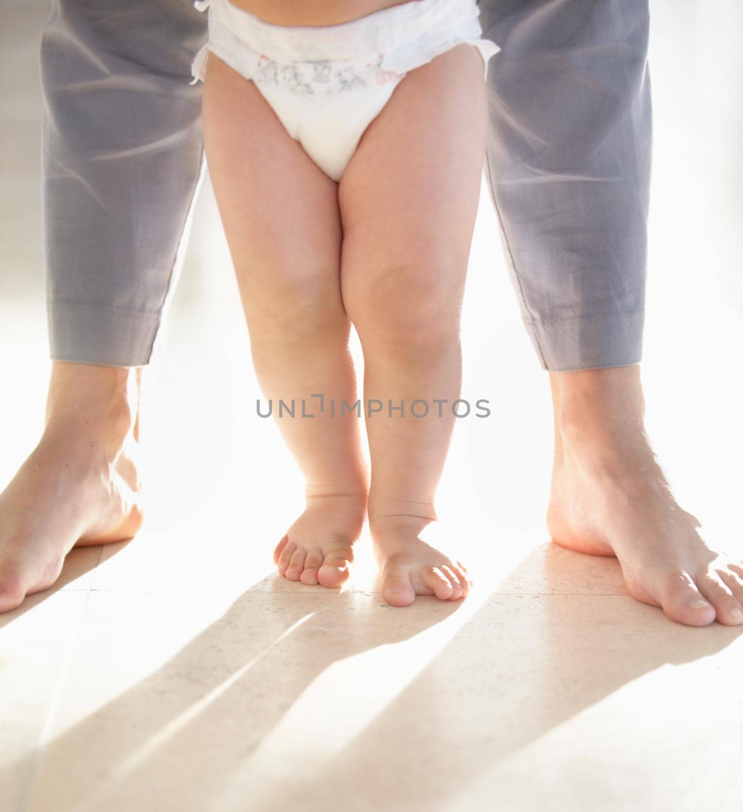 Cropped image of the legs of a baby walking with a parent supporting from behind.